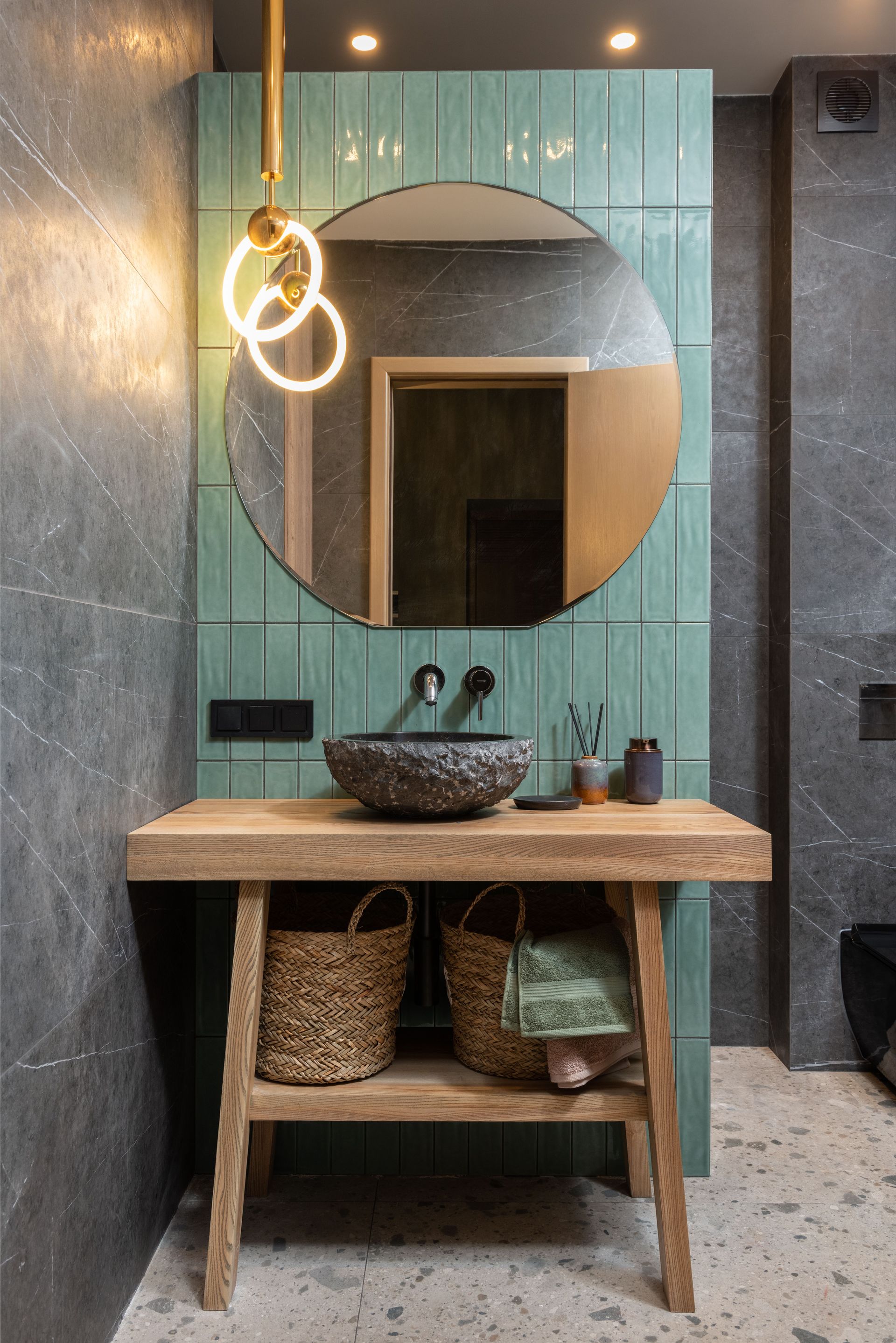 Moderne badkamermeubel met houten blad, ronde spiegel, gevlochten mand en hanglamp tegen een groenblauwe tegelwand