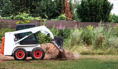 White skid-steer loader digging dirt in a grassy yard near a fence and bushes