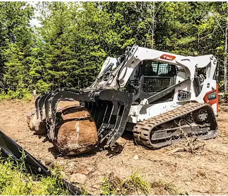 White compact loader with grapple moving a large log on a dirt trail in a forest.