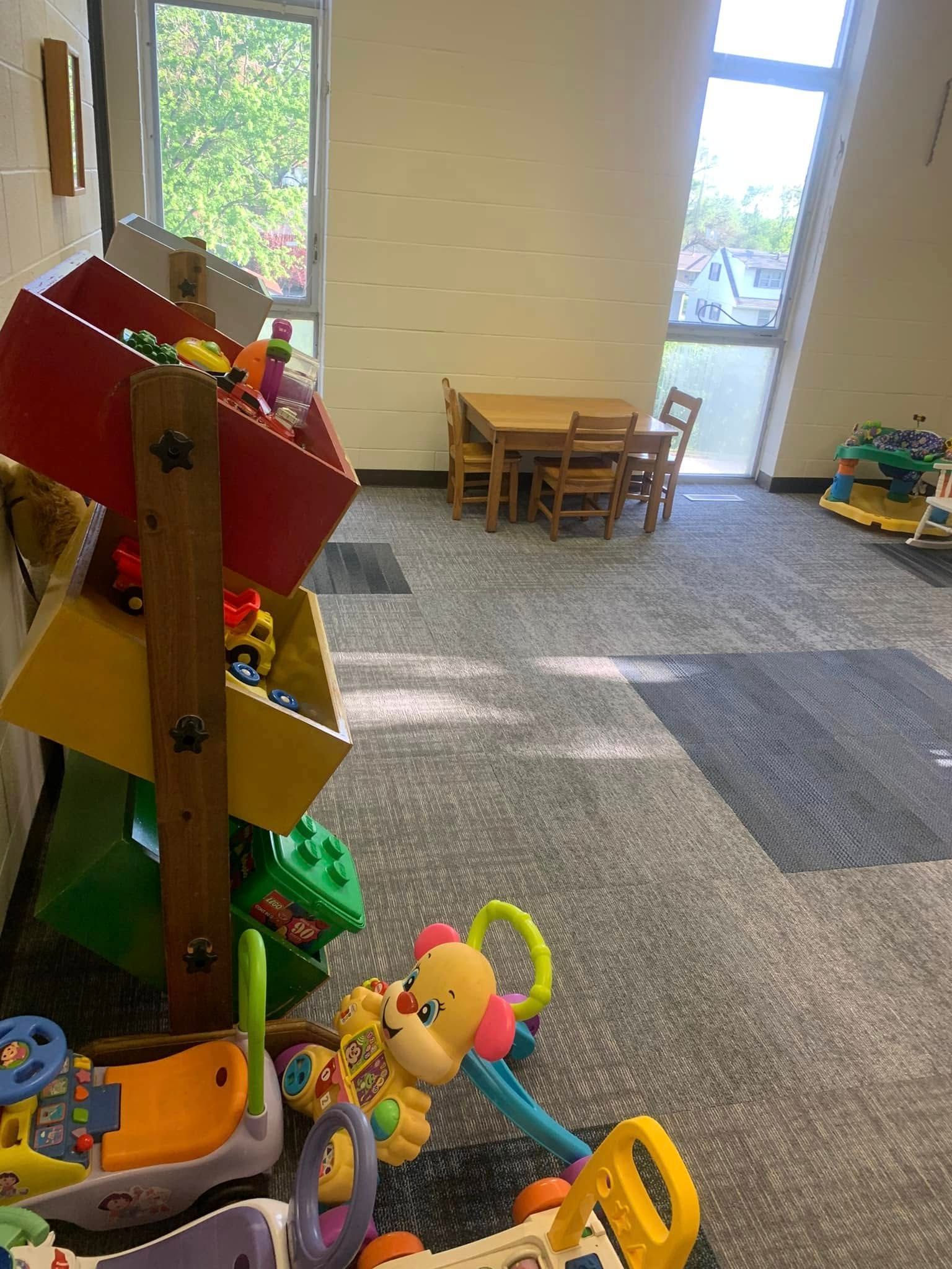 Child’s playroom with toys in the foreground, small table and chairs by bright windows, and a carpeted floor.