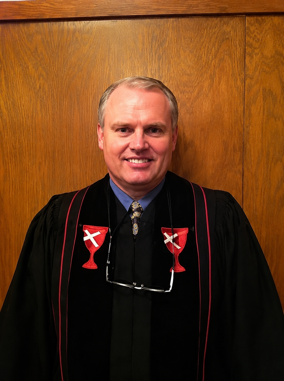 Man in black robe with red trim smiling against a wood-paneled wall