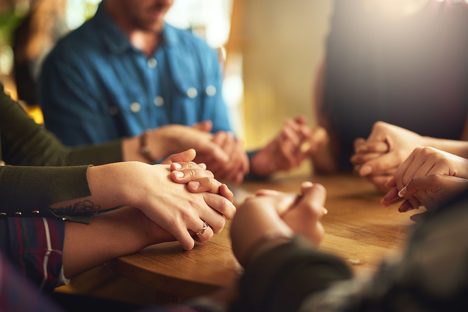 Several hands are held together in prayer over a Bible resting on a wooden table.