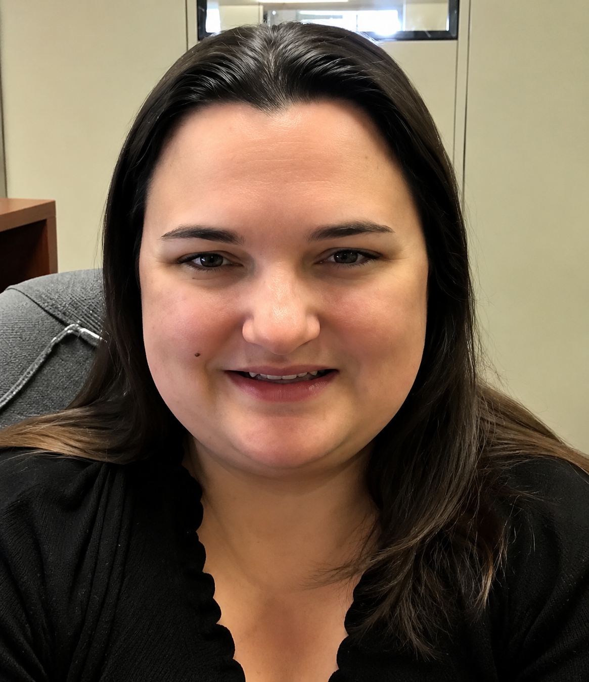 Smiling person with dark hair in a black top, seated in an office setting.