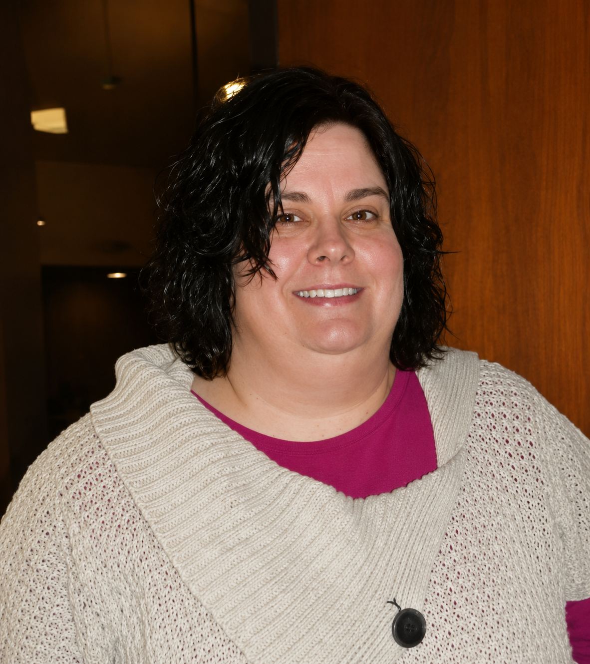 Smiling person with dark hair in a cream sweater over a magenta shirt, standing indoors.
