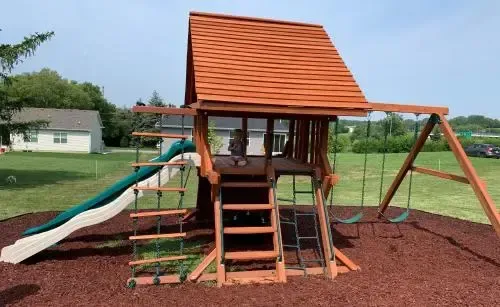 Children’s playground with a wooden playset, orange roof, slide, ladder, and swing on a grassy lawn