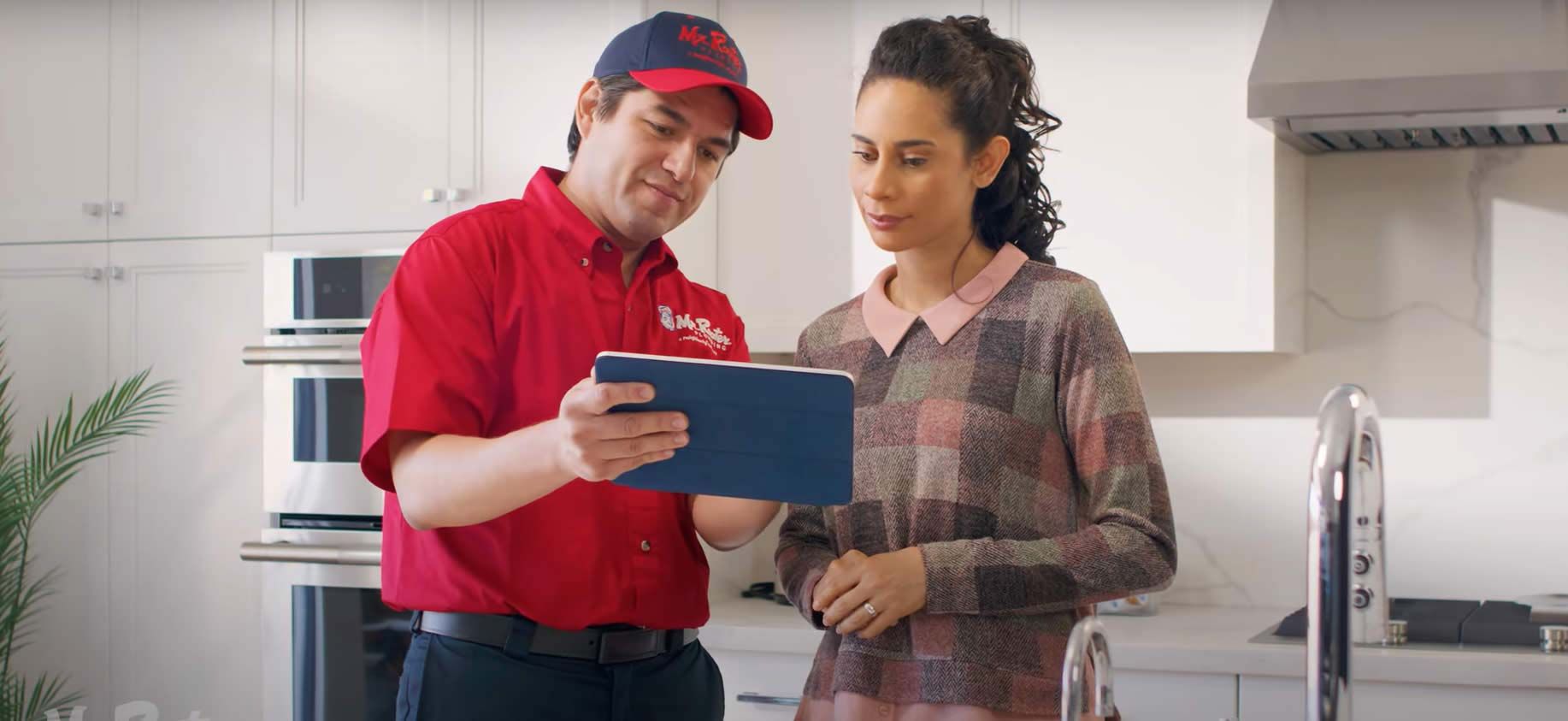 A professional wearing a red uniform and cap shows a tablet screen to a person while standing in a home.