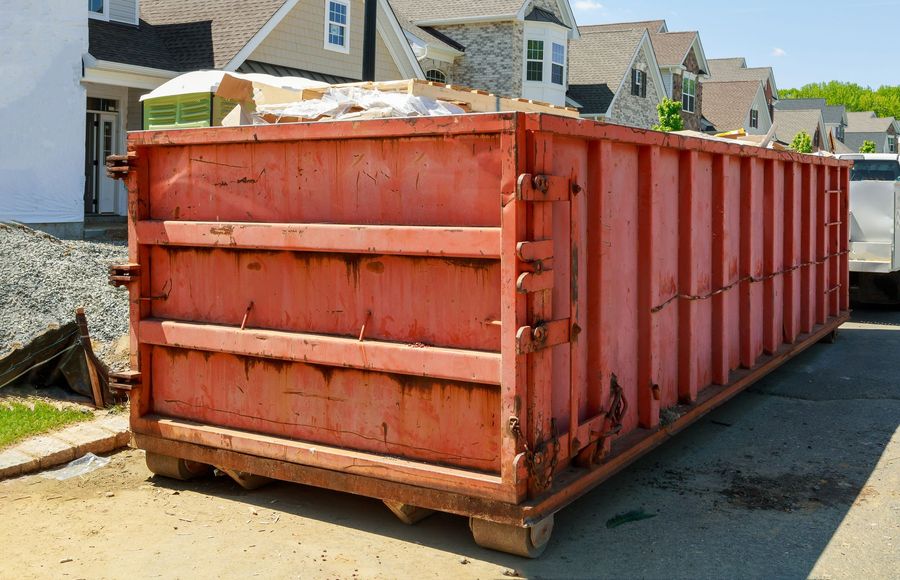 Red construction dumpster on a street beside houses, loaded with debris