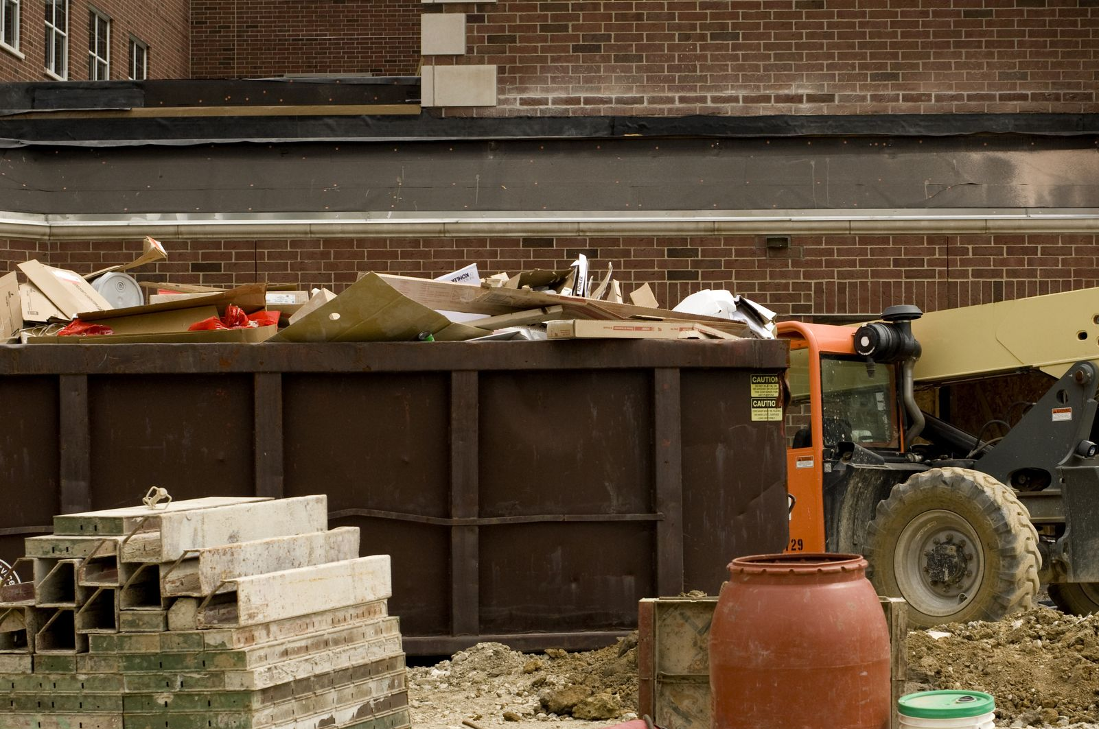 Construction debris in a dumpster beside a wheelbarrow, cinder blocks, and a brick wall.
