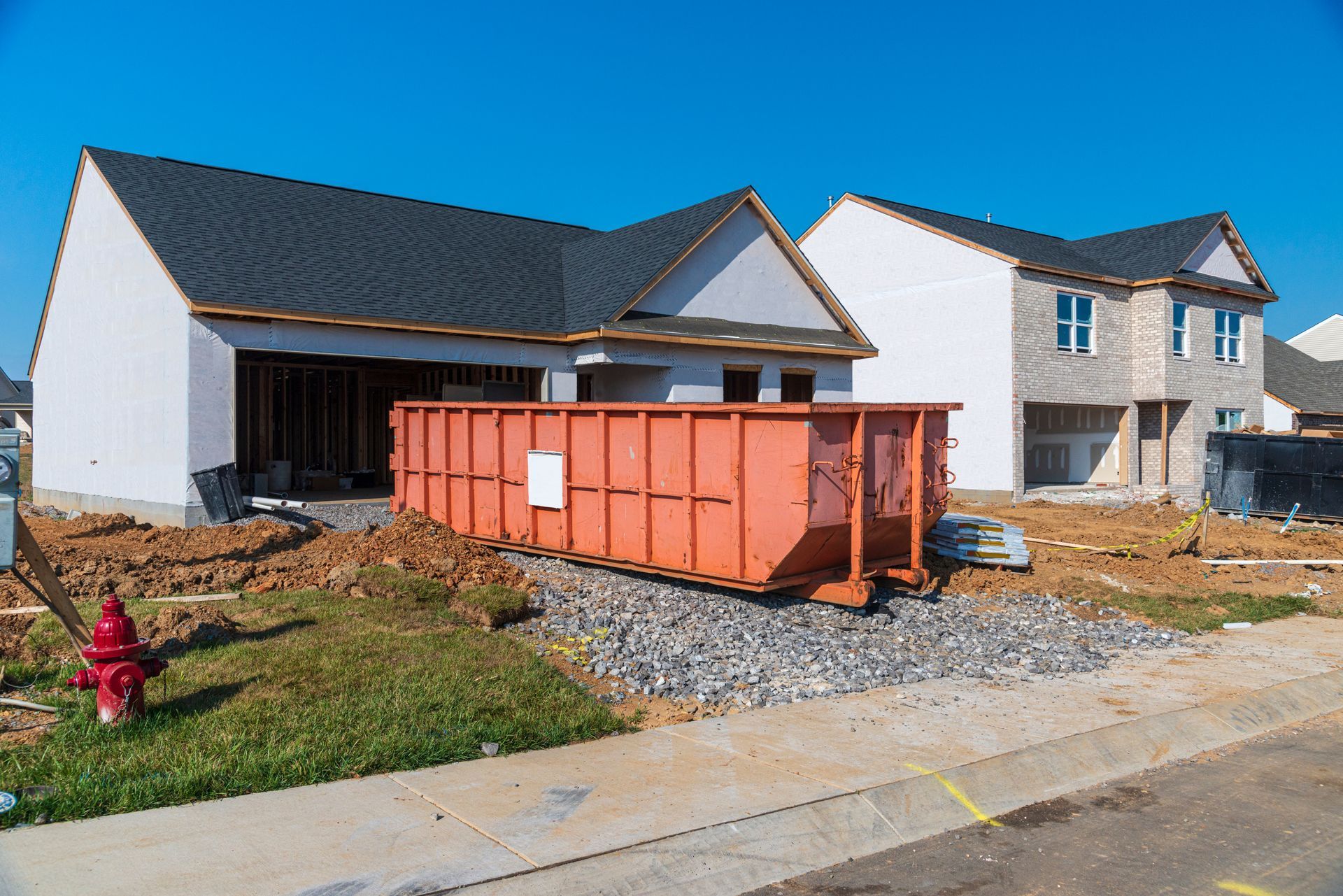 New homes under construction with an orange dumpster, gravel pile, and fire hydrant by the curb.