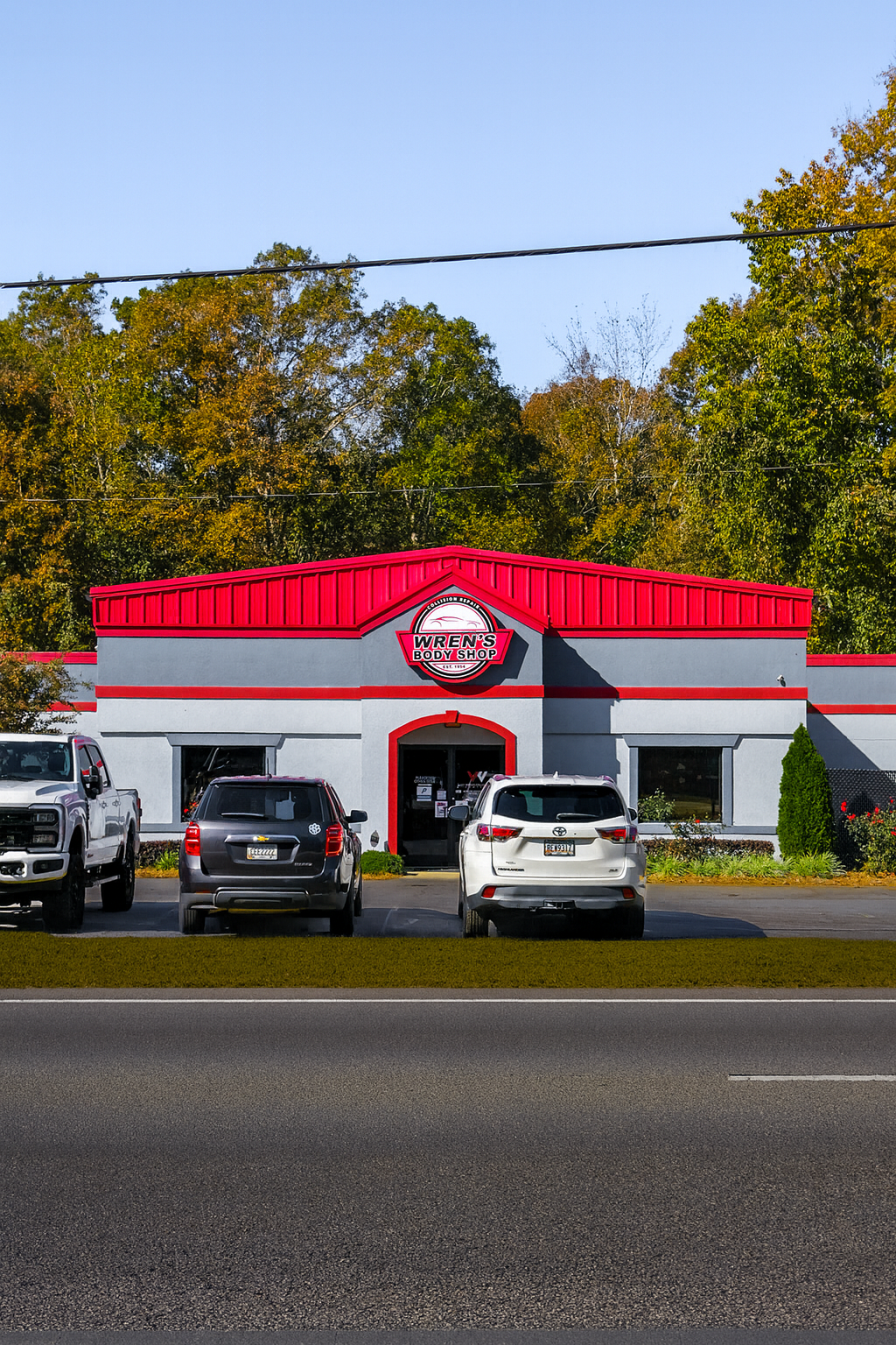 A storefront with a red roof and gray walls. Cars are parked in front. Fall foliage in the background.