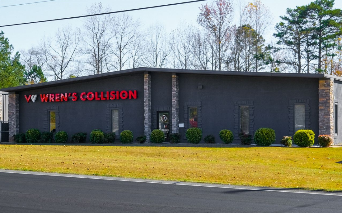 Wren's Collision building exterior. Dark gray with red sign, stone accents. Grass lawn, trees in background.