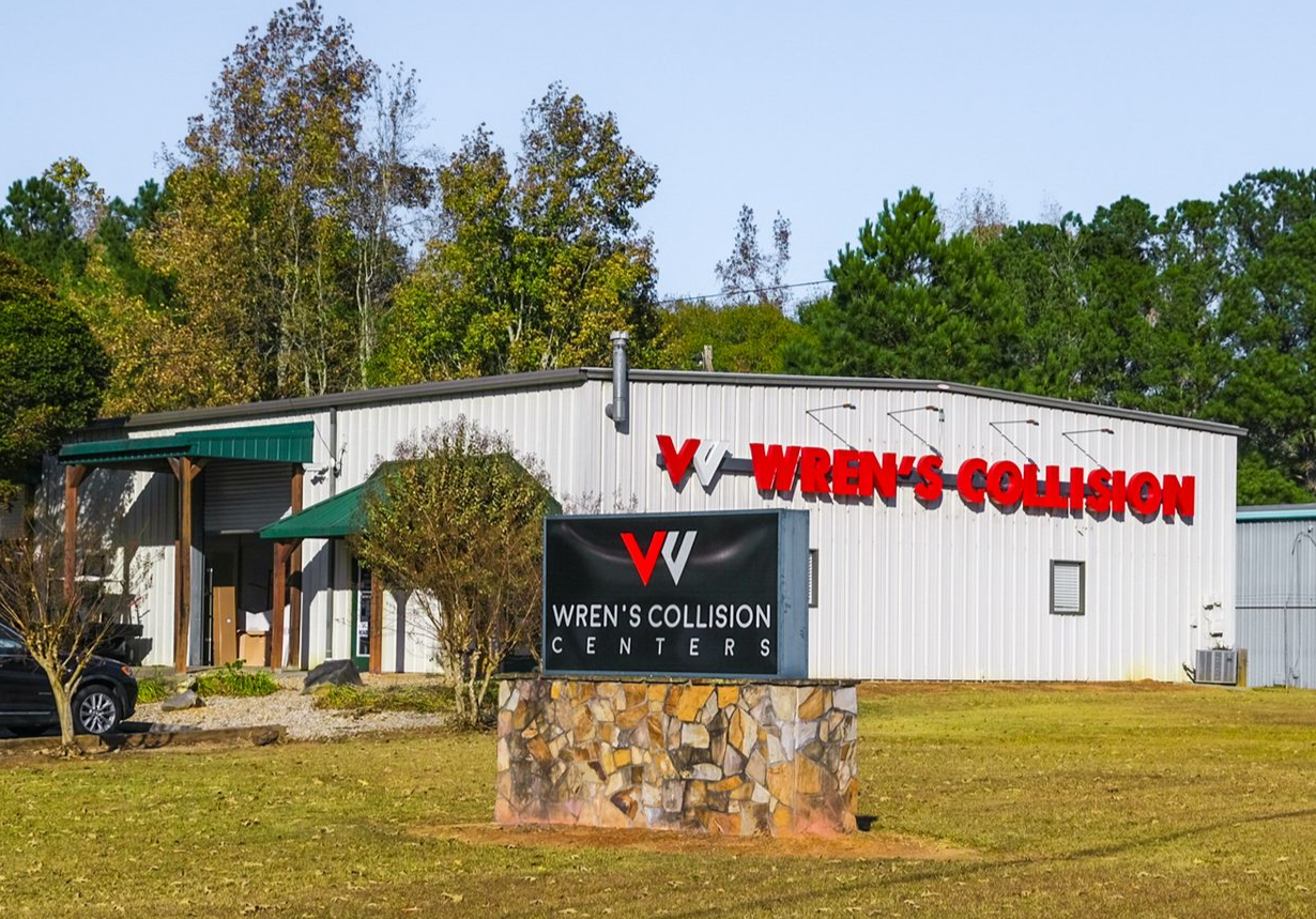 Wren's Collision Center building with a sign on a grassy area. Red and black sign, trees in background.