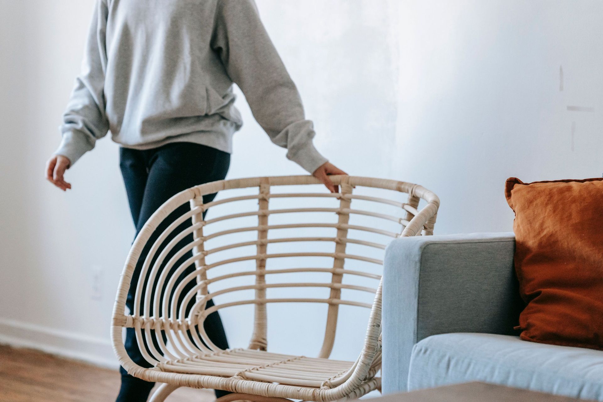 A person wearing a grey sweatshirt moving a round rattan chair inside a brightly lit living room space.