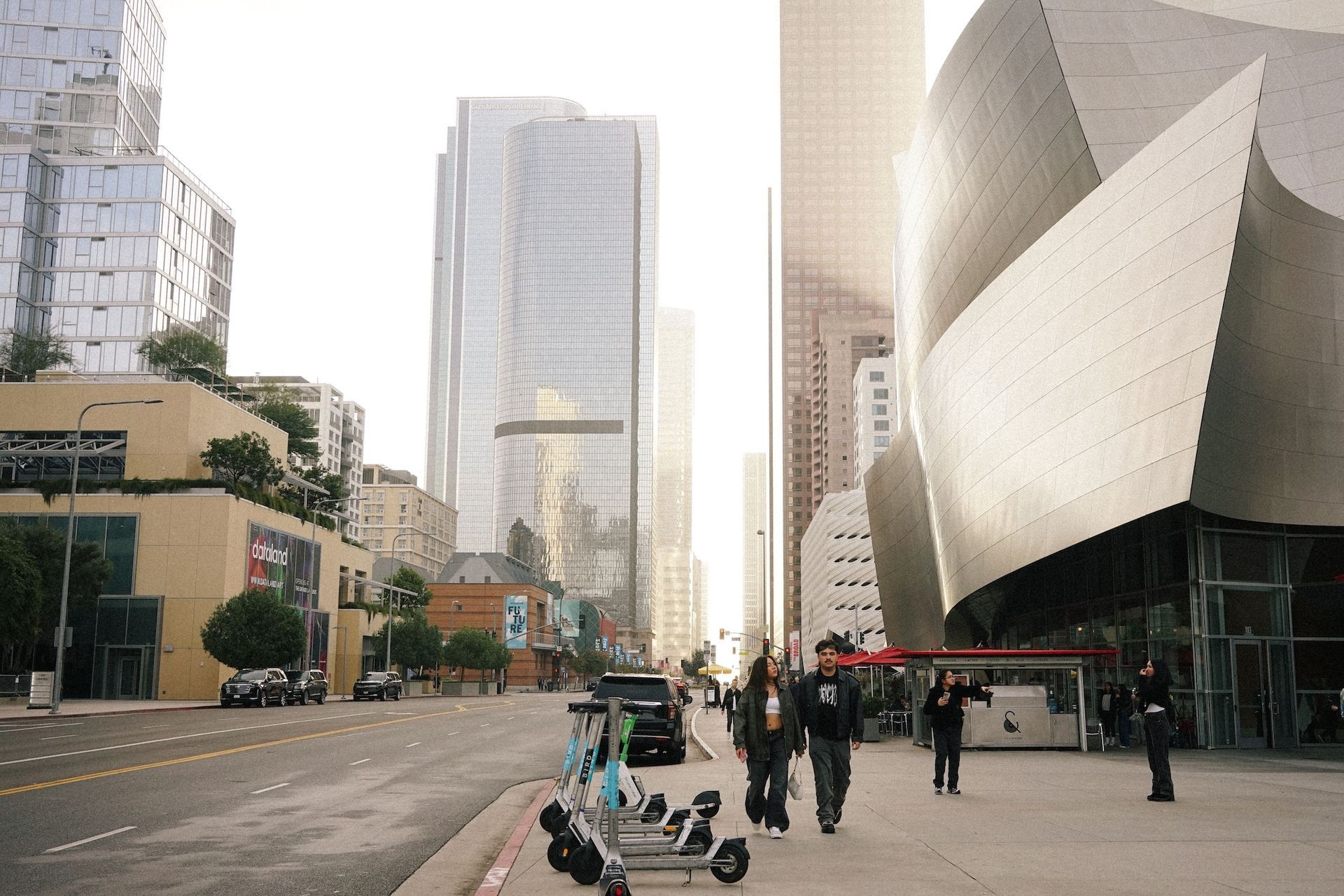 Two people walking past parked electric scooters and a curved metallic building standing along a bright city street.