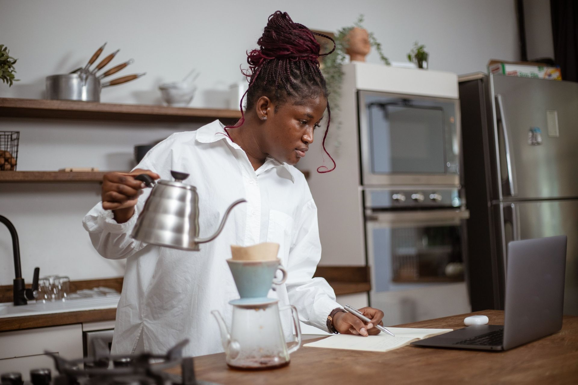A woman pouring water from a gooseneck kettle into a pour-over filter while writing notes next to a laptop.