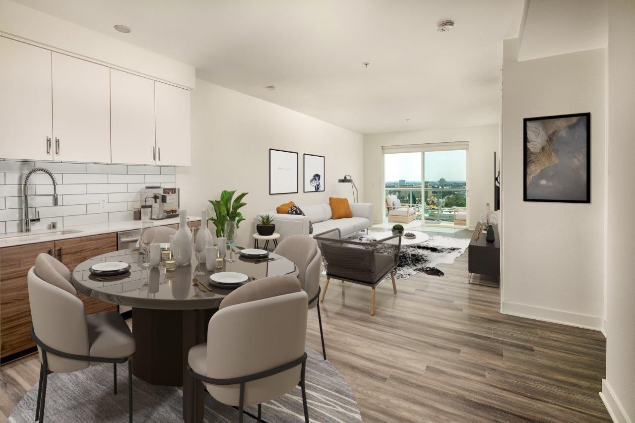 An open-concept apartment layout showing a round dining table, kitchen cabinets, living area, and a balcony door.