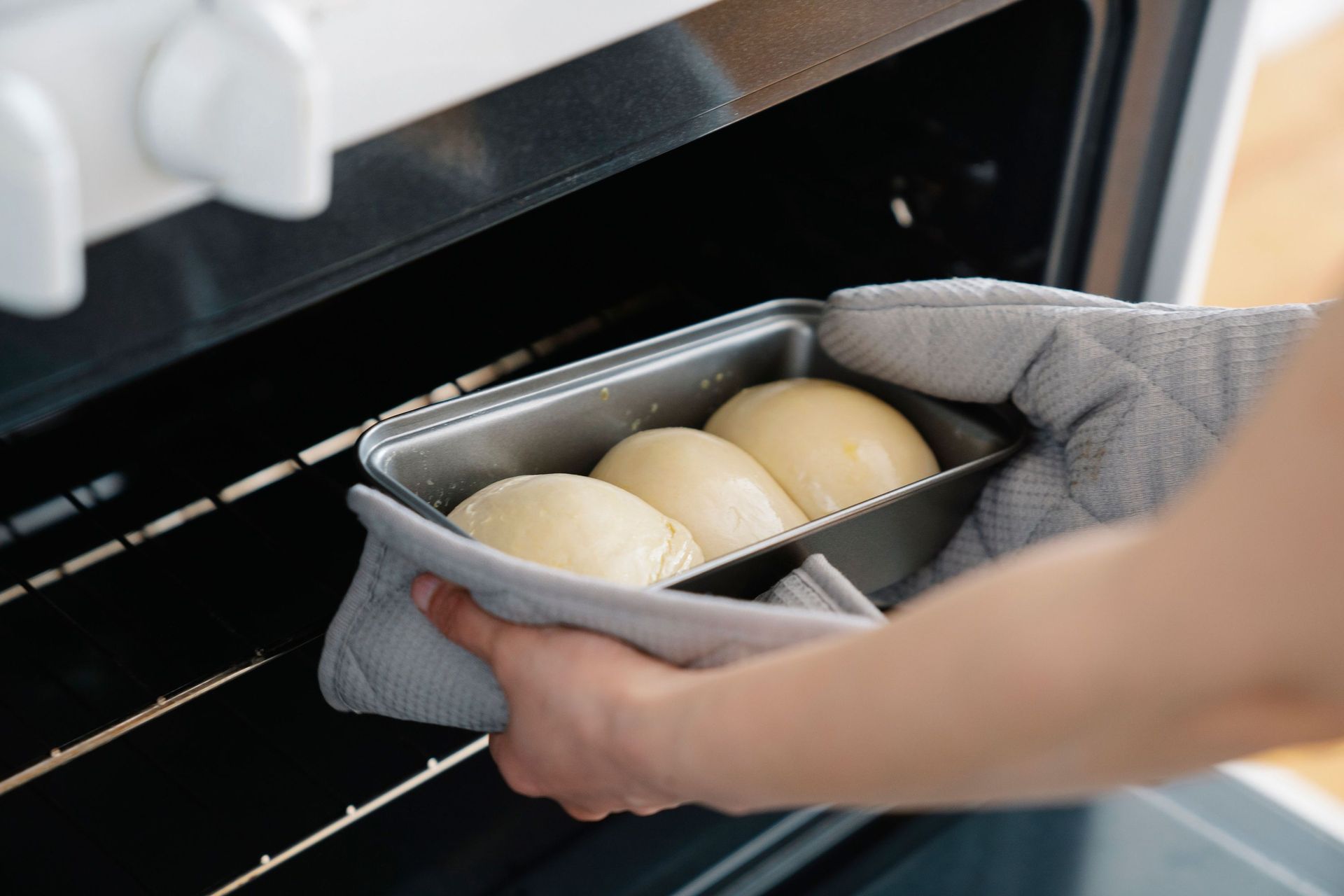 A person wearing oven mitts carefully placing a loaf pan of unbaked bread dough into a hot kitchen oven.