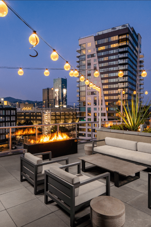 Rooftop patio with modern furniture, fire pit, and string lights, overlooking city skyline at dusk.