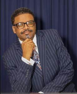 Man in a navy pinstripe suit and glasses poses thoughtfully against a dark curtain backdrop.