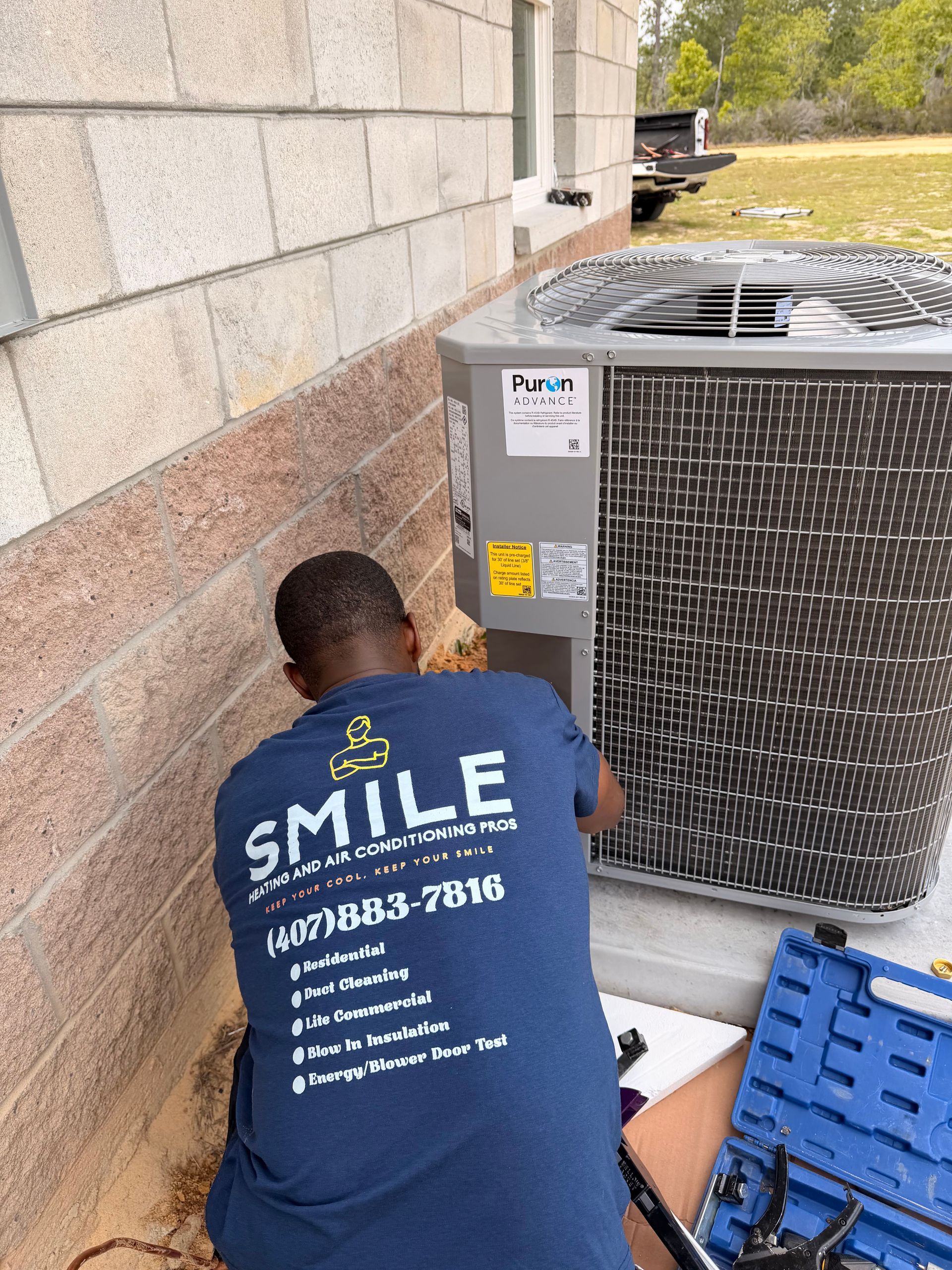 Technician kneeling by an HVAC unit beside a building wall, working on equipment with tools nearby.