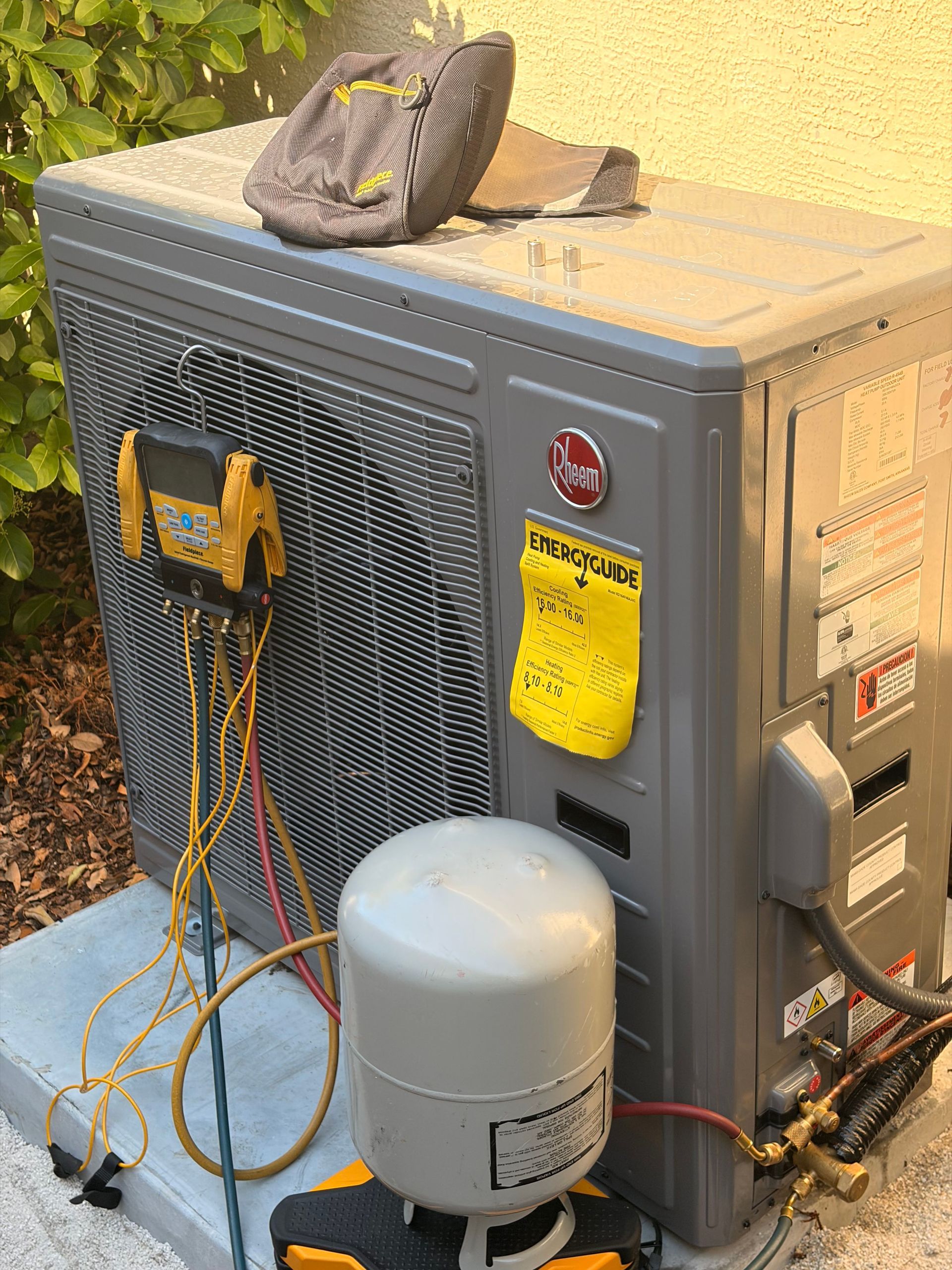 Outdoor HVAC unit with maintenance tools and hoses attached, next to a white tank and covered by leaves