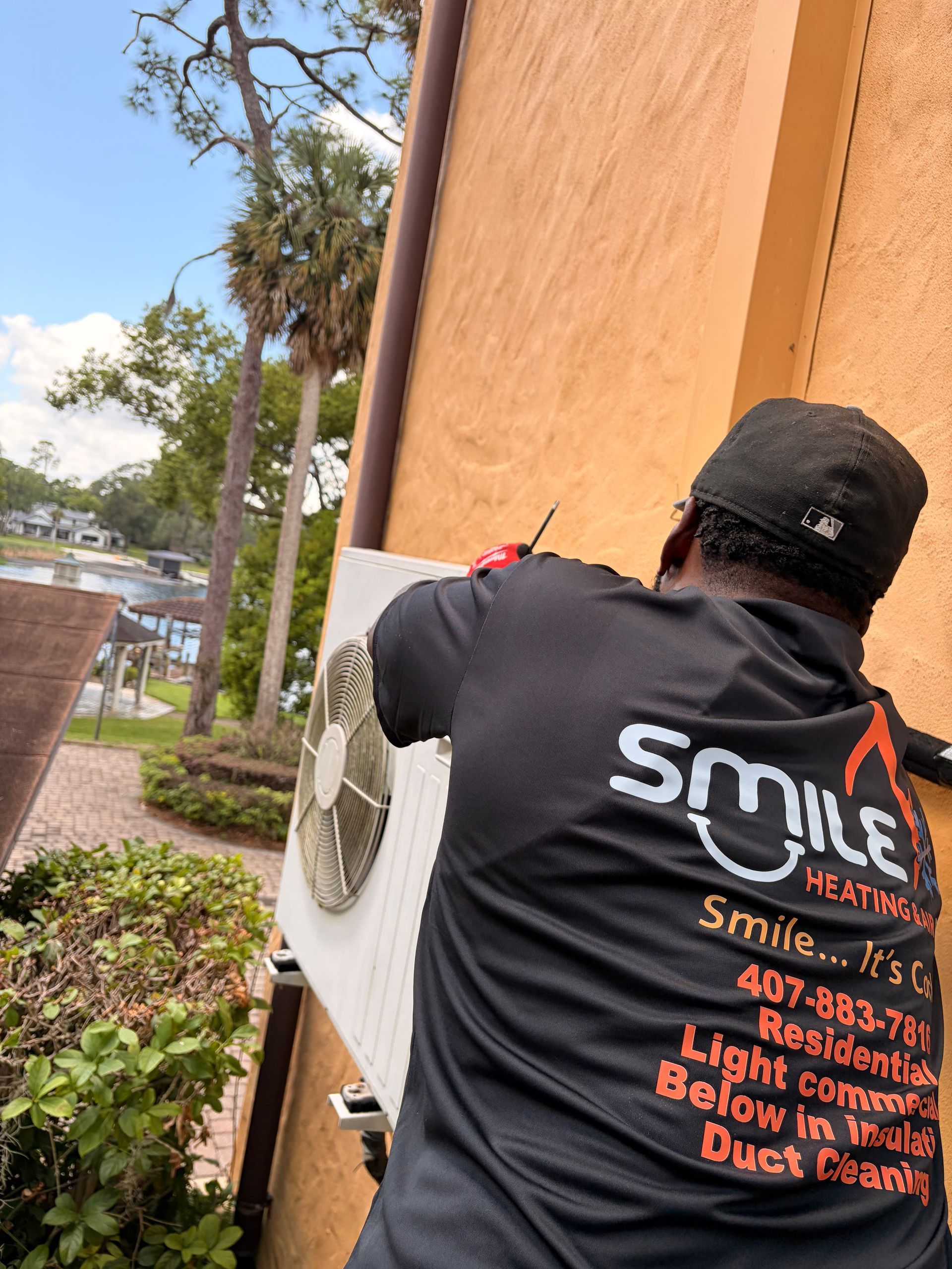 Technician installing a wall-mounted air conditioner beside a building, wearing a black Smile Services shirt.