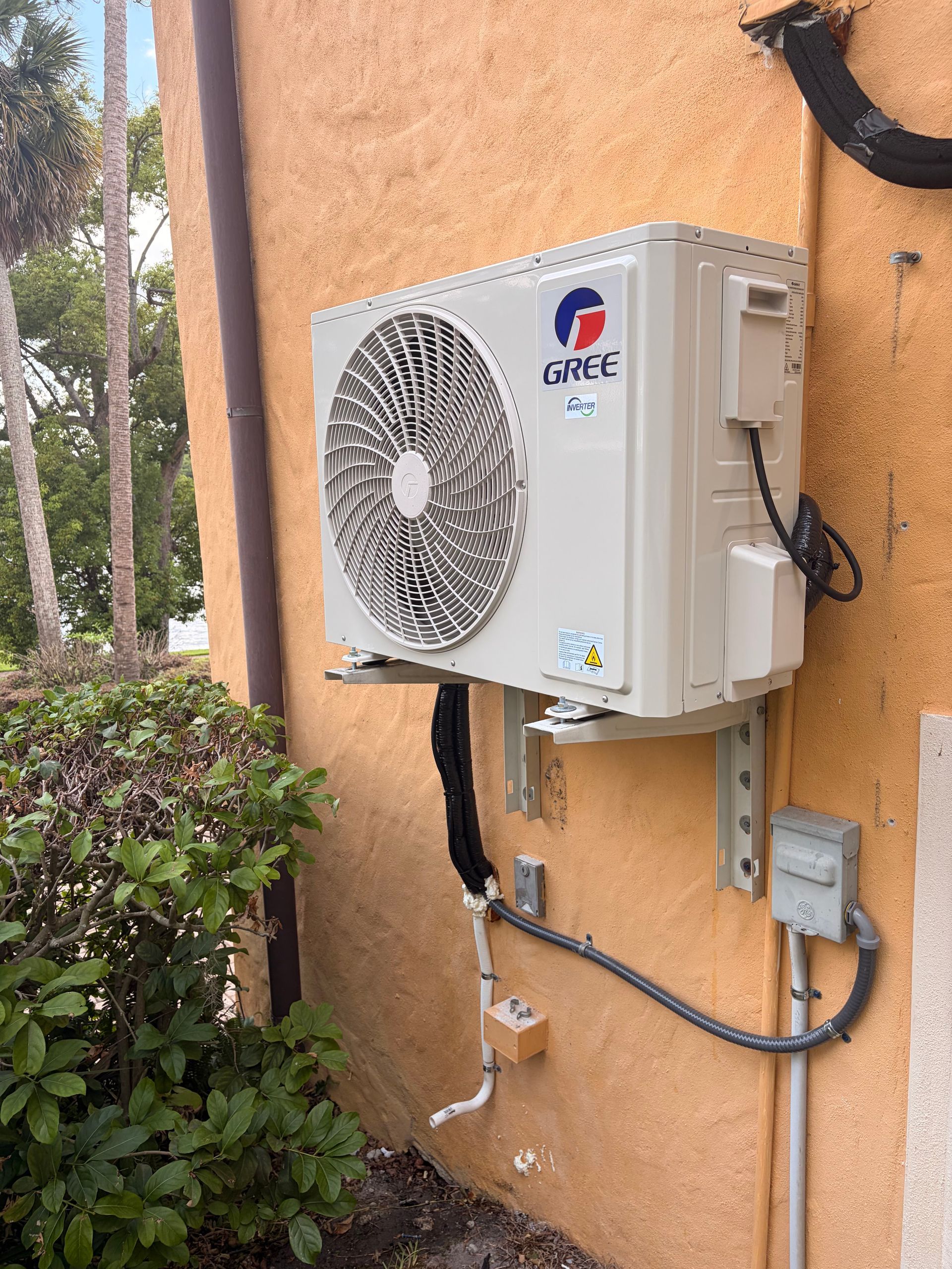 Wall-mounted outdoor air conditioner unit on a peach stucco wall with plants nearby.