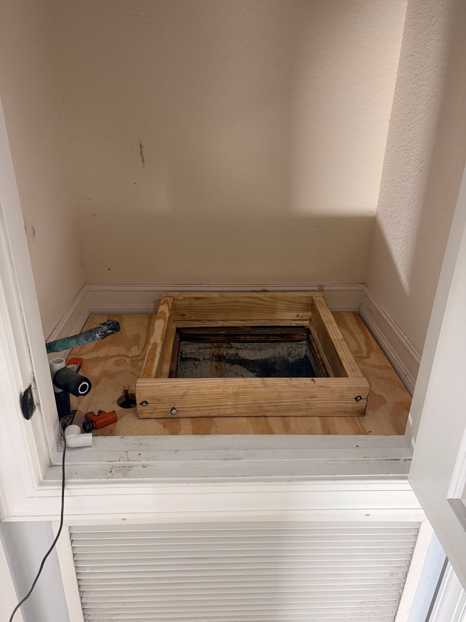 Open closet with exposed floor opening framed by wood, tools on the left, and white louvered doors below.