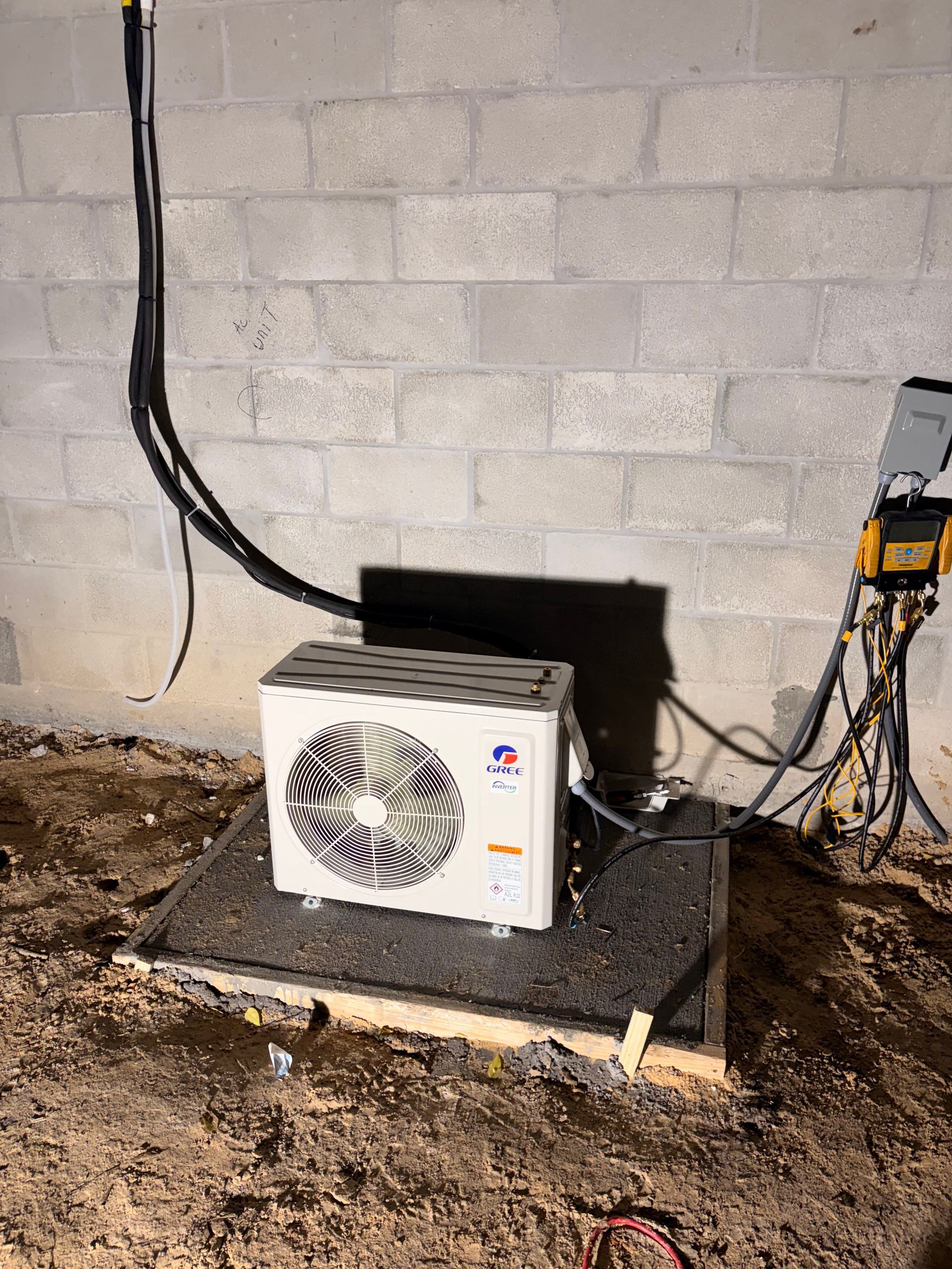 Outdoor air conditioner unit beside a cinderblock wall with exposed black cables and electrical box.