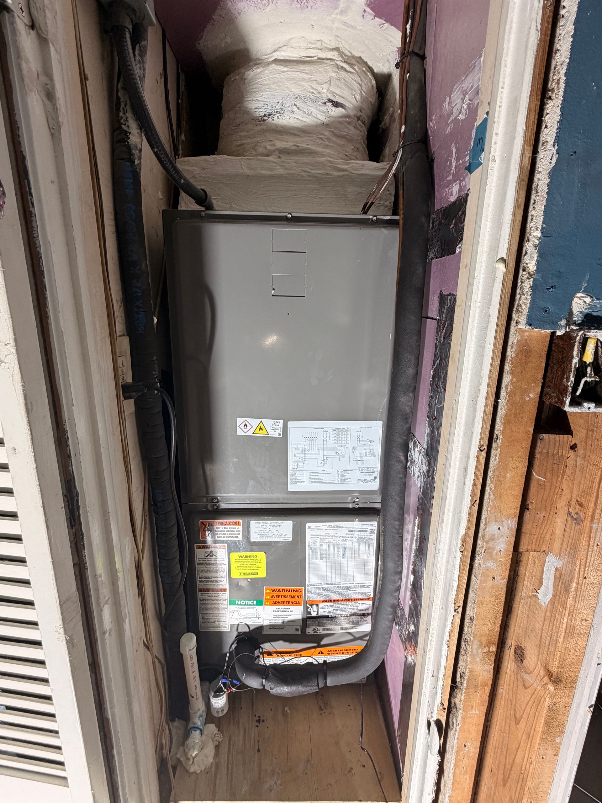 Gray furnace unit in a narrow utility closet with exposed walls and a duct opening above.