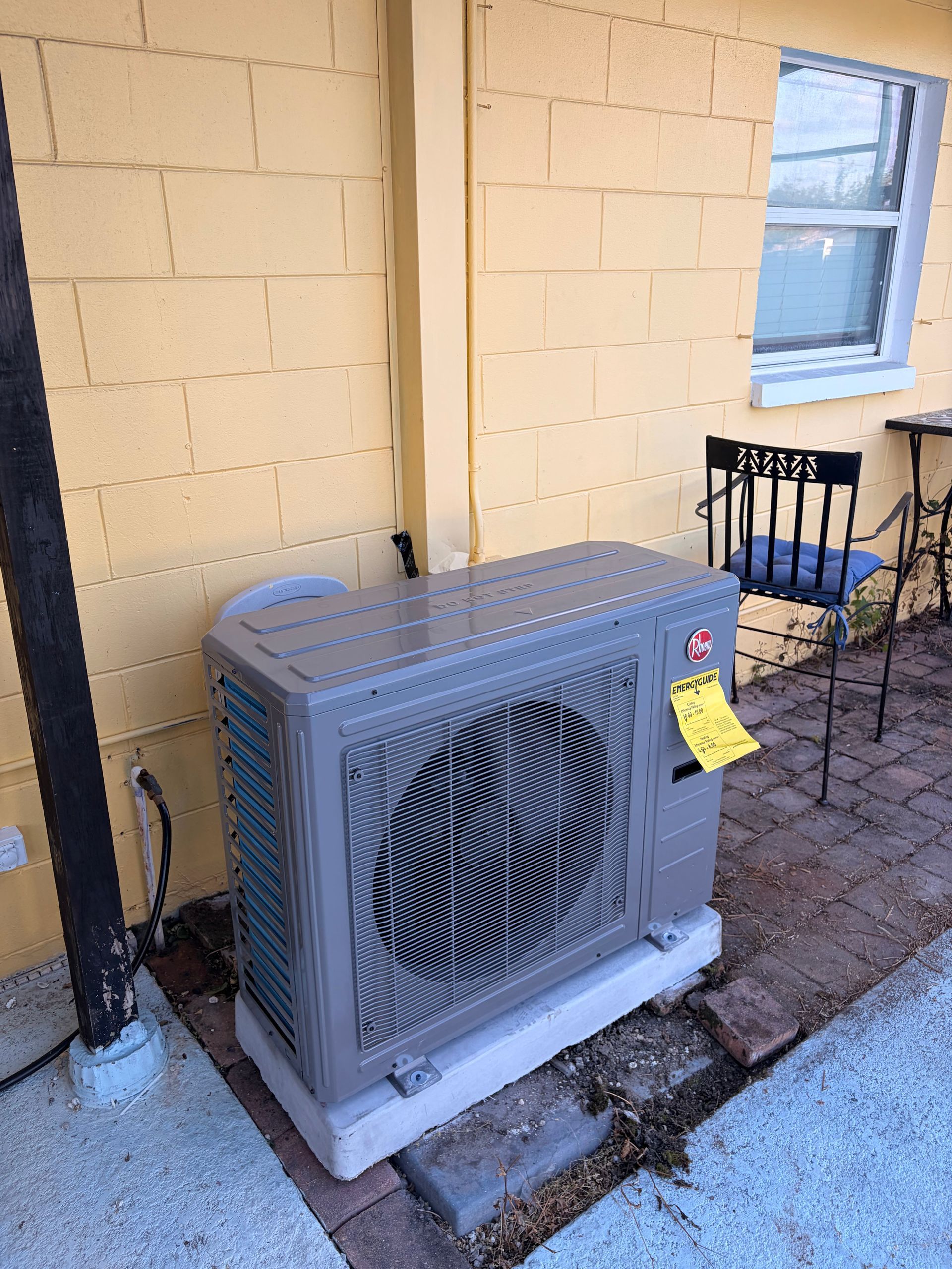 Outdoor HVAC unit beside a yellow building wall, near a window and metal bench on a brick patio.