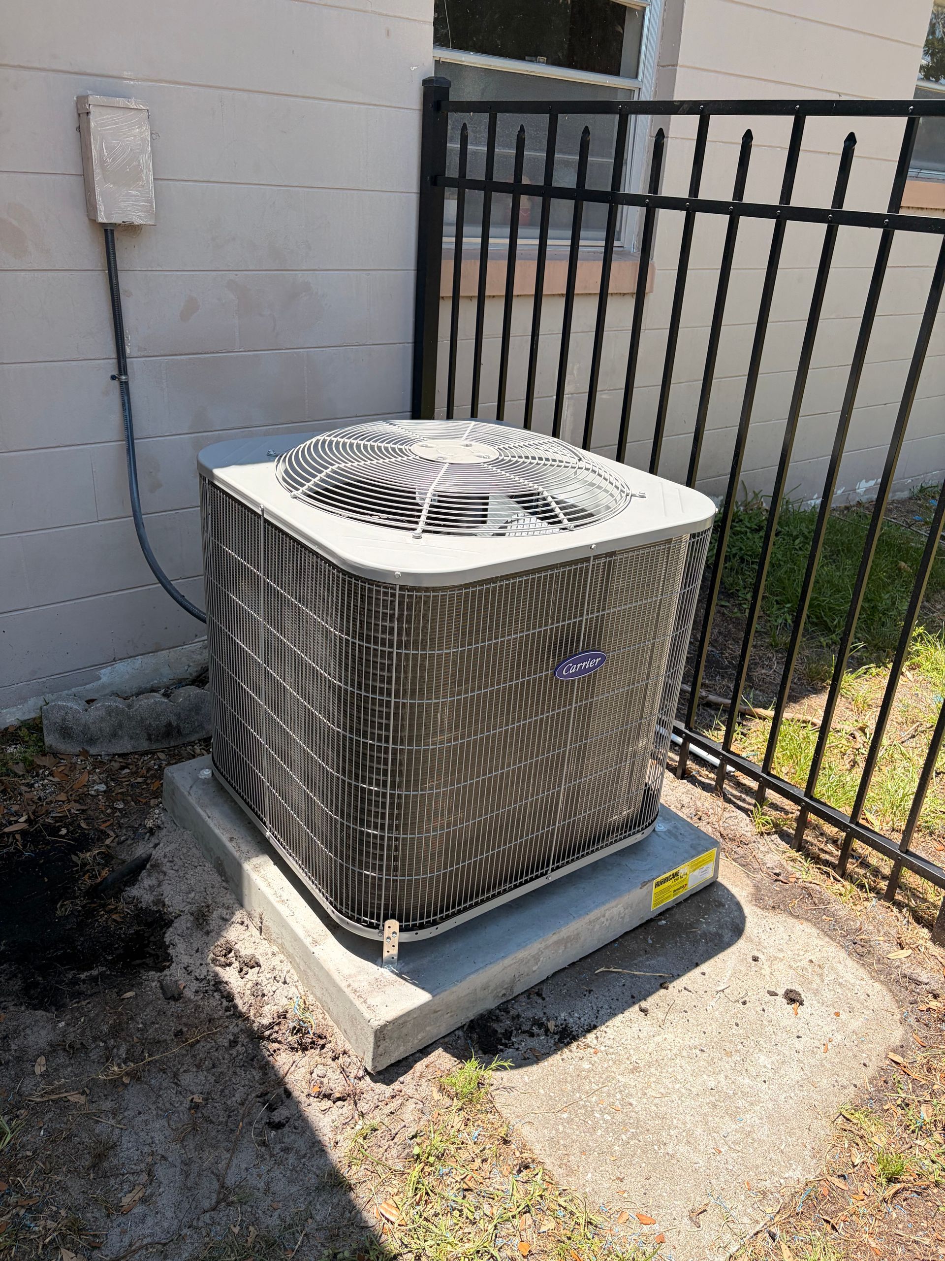 Outdoor air conditioner unit beside a cinder block wall and black metal fence on a concrete pad