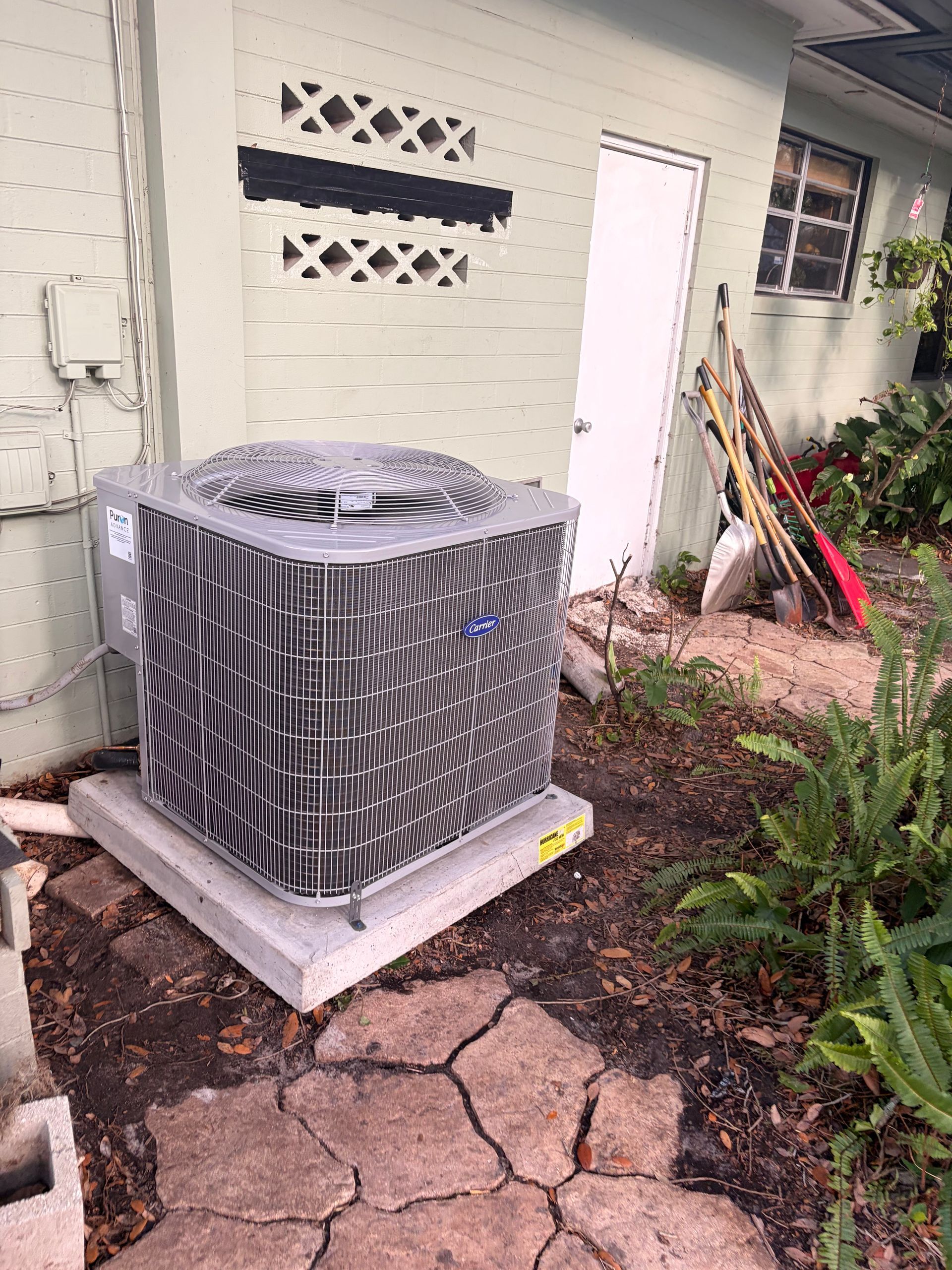 Outdoor AC unit beside a cracked walkway and house wall, with garden tools leaning nearby.