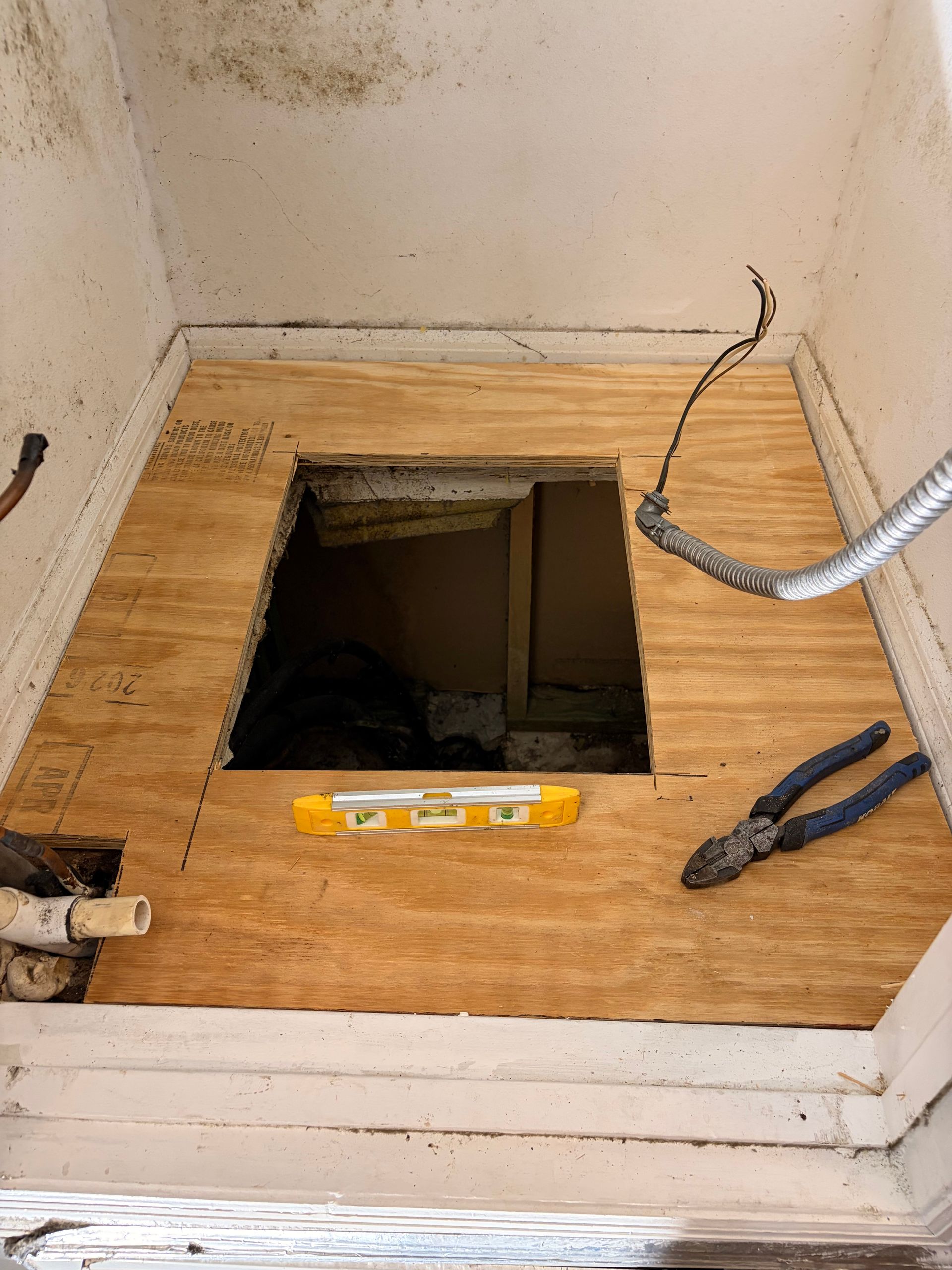 Small unfinished room floor with square opening, exposed wiring, and tools on a plywood surface