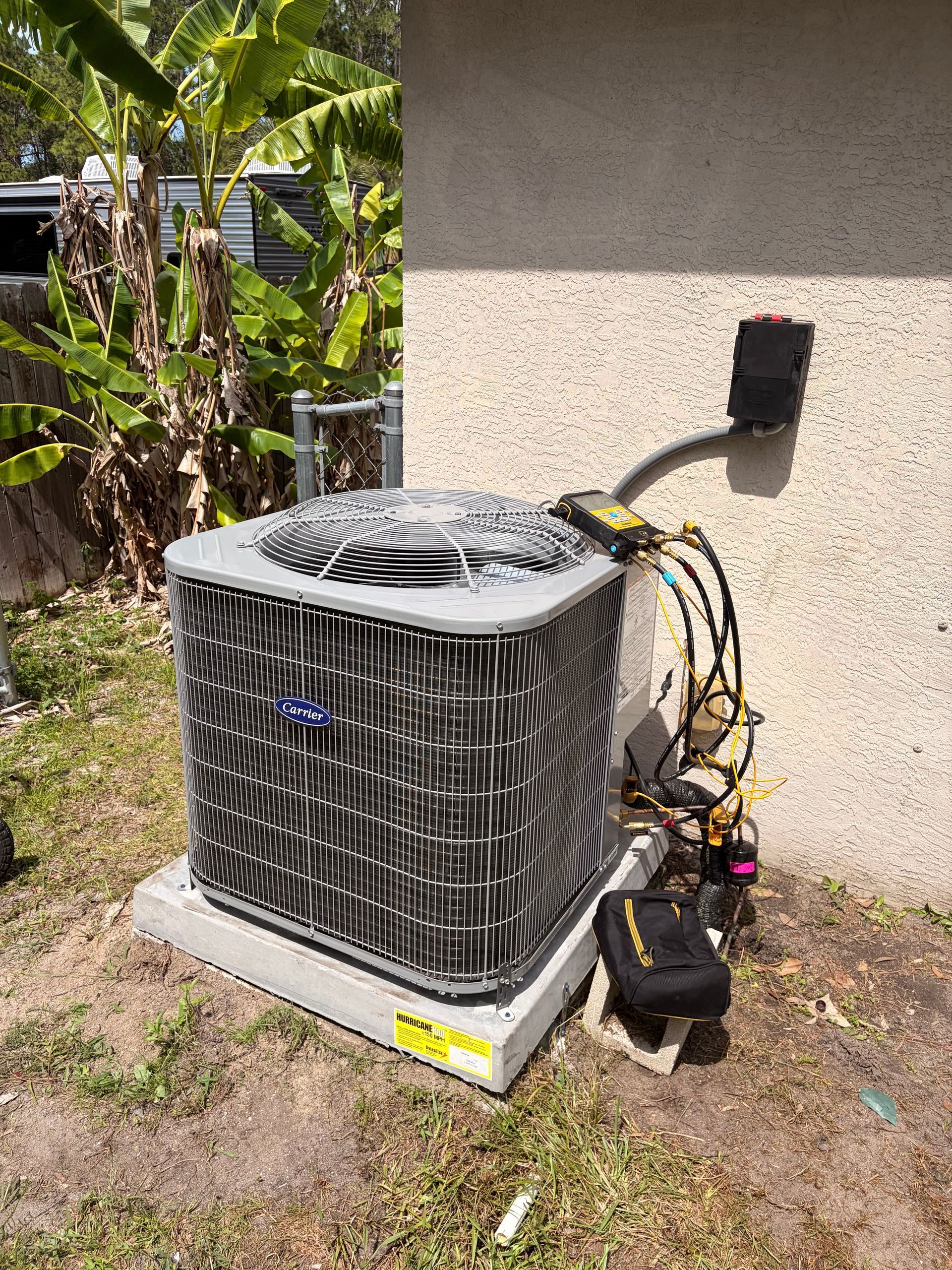 Outdoor air conditioner unit beside a house wall, with exposed wires and hoses on a gravel patch.
