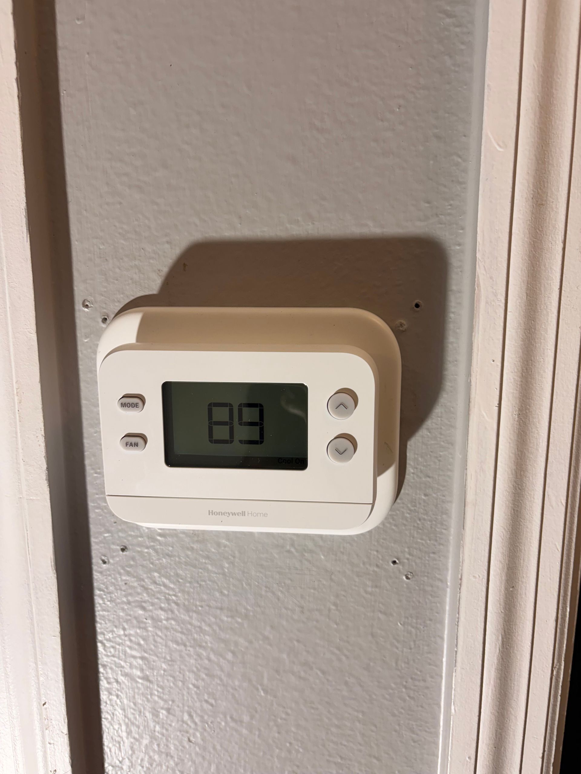 Wall-mounted digital thermostat with display reading 19 on a beige wall beside a doorway