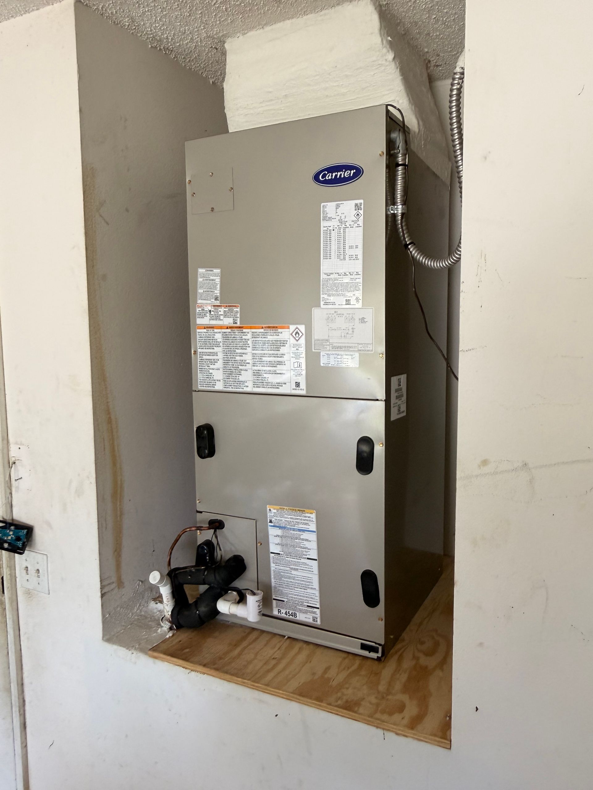 HVAC furnace in a recessed wall alcove with exposed wiring and pipes, gray metal unit on a plywood shelf