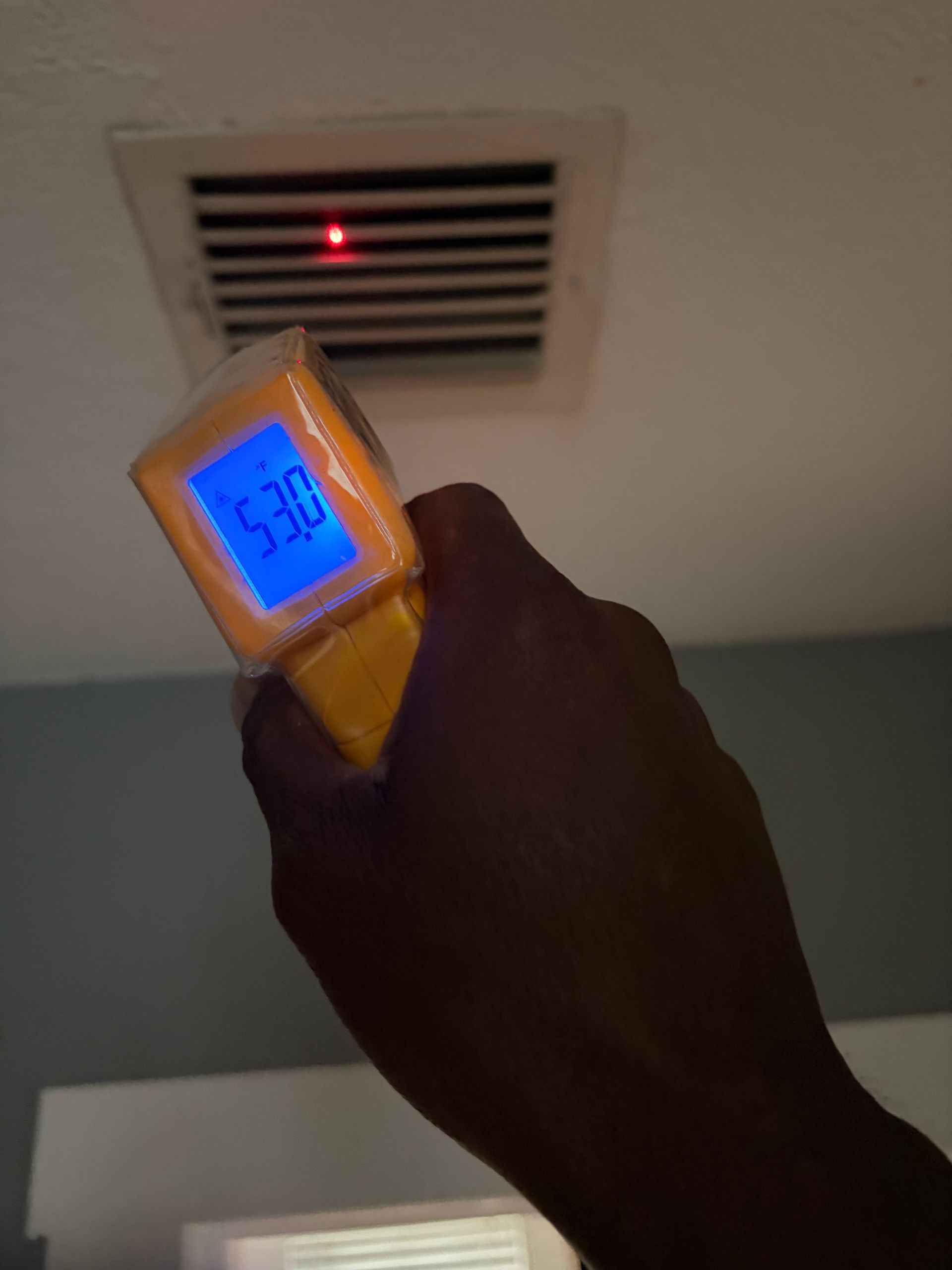 Hand holding a yellow infrared thermometer aimed at a ceiling vent with a red light.