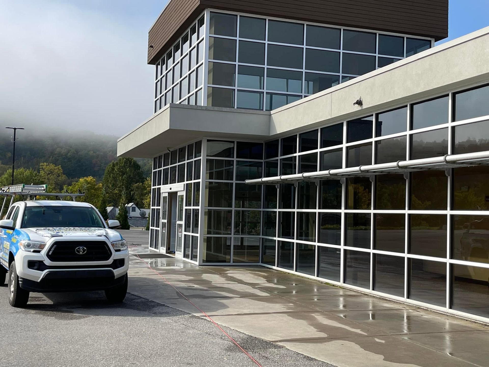 White SUV parked outside a glass-front building on a wet pavement, with mountains in the background