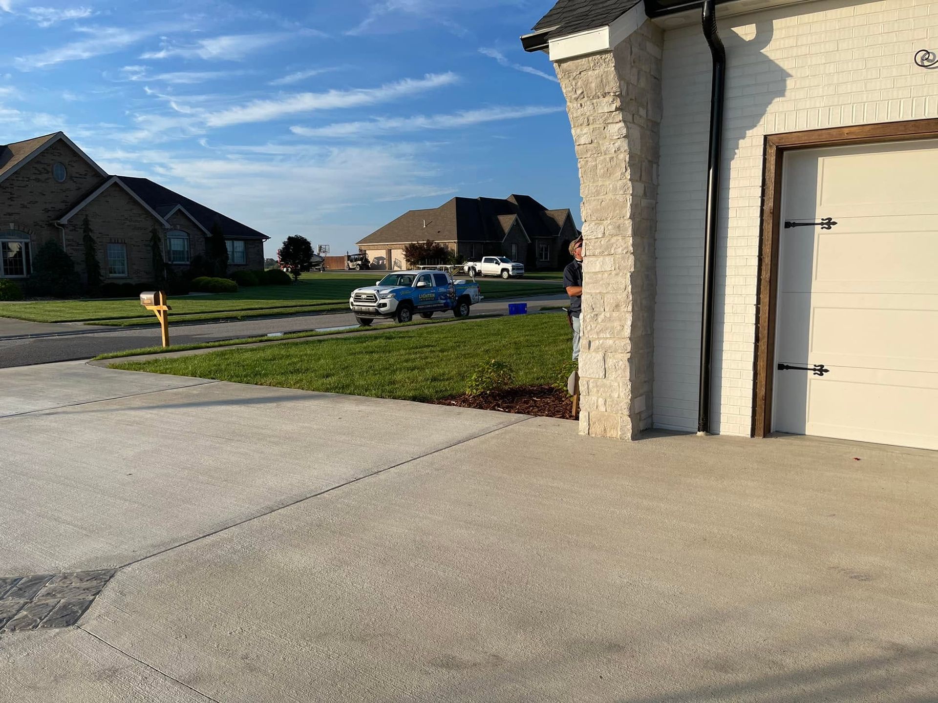 Suburban street view with driveway, garage wall, lawn, and houses under a blue sky.