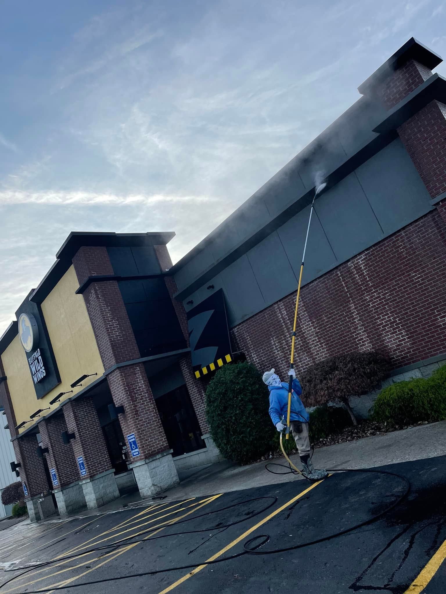 A worker uses a long pole to inspect or clean a building roof near a brick-and-stucco storefront.