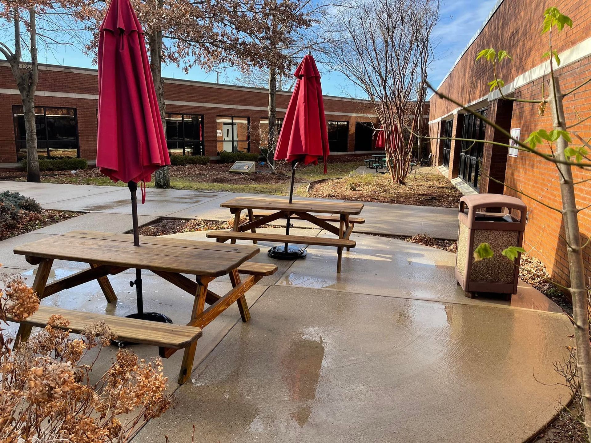 Outdoor courtyard with wet picnic tables, red umbrellas, and a trash bin near brick buildings.