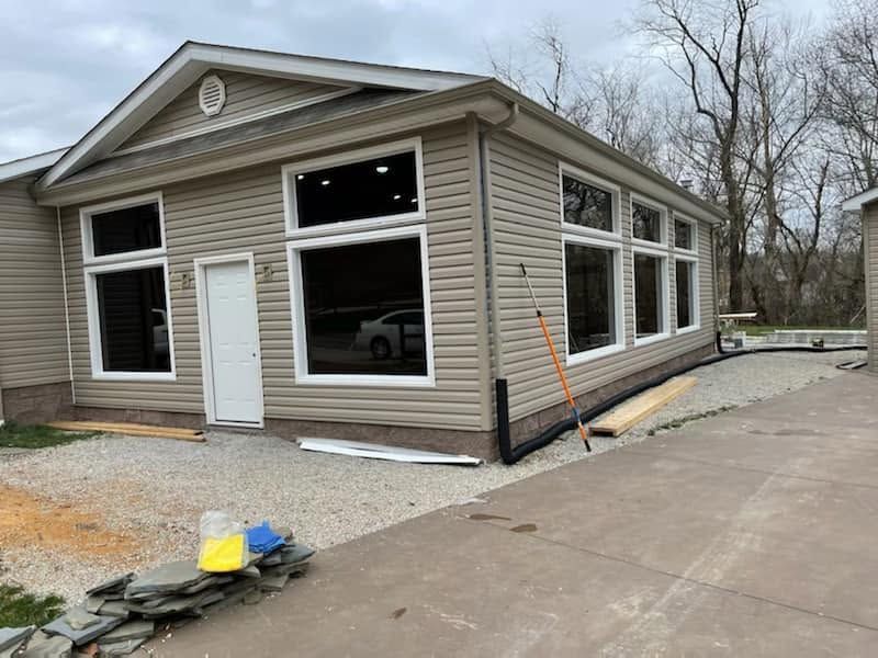 Tan house under construction with large windows, gravel, driveway, and siding tools nearby