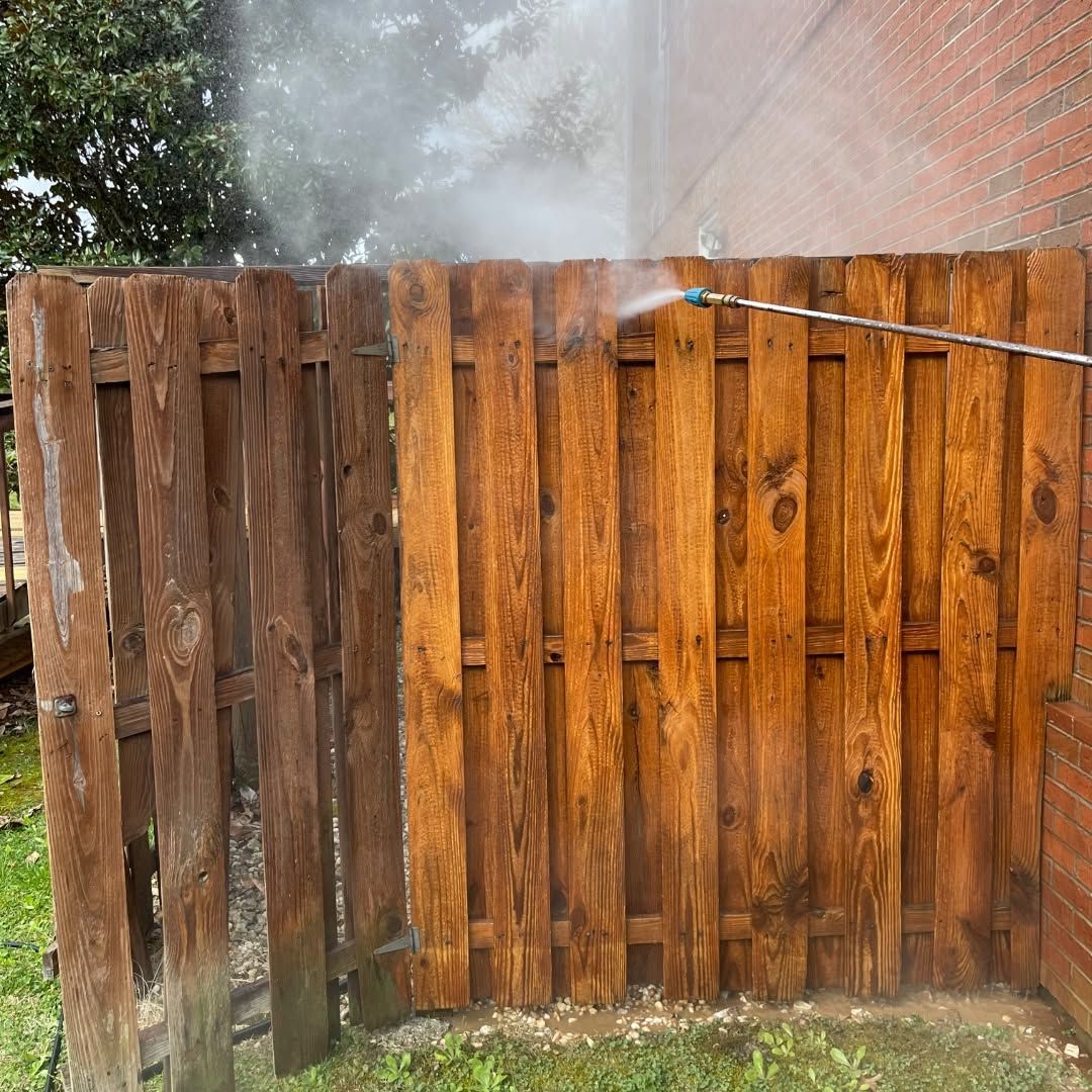 Wooden fence being spray-washed with water mist in a backyard, beside a brick wall.