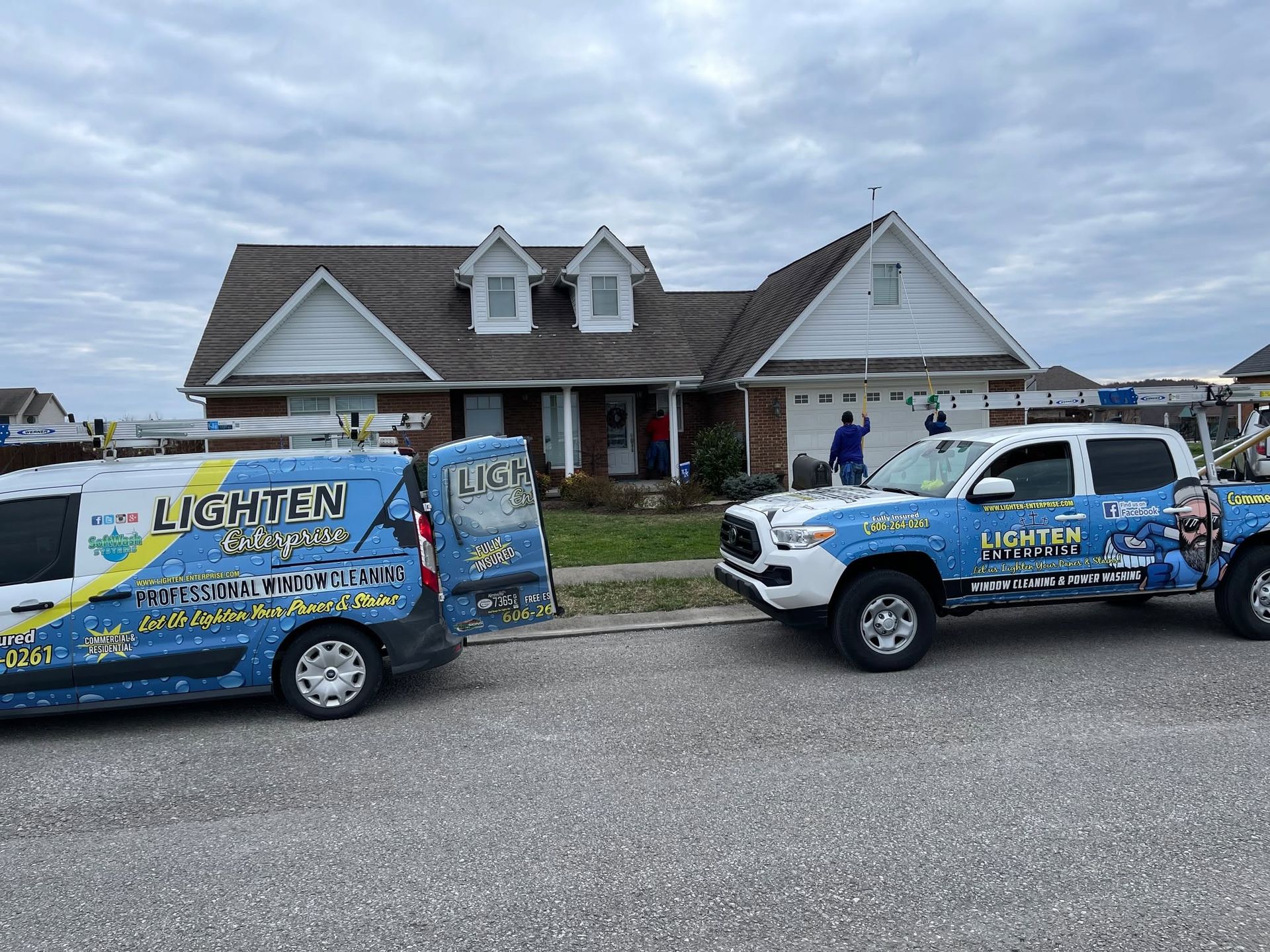 Two blue utility vans parked in front of a house under repair, with a ladder on the roof and overcast sky.