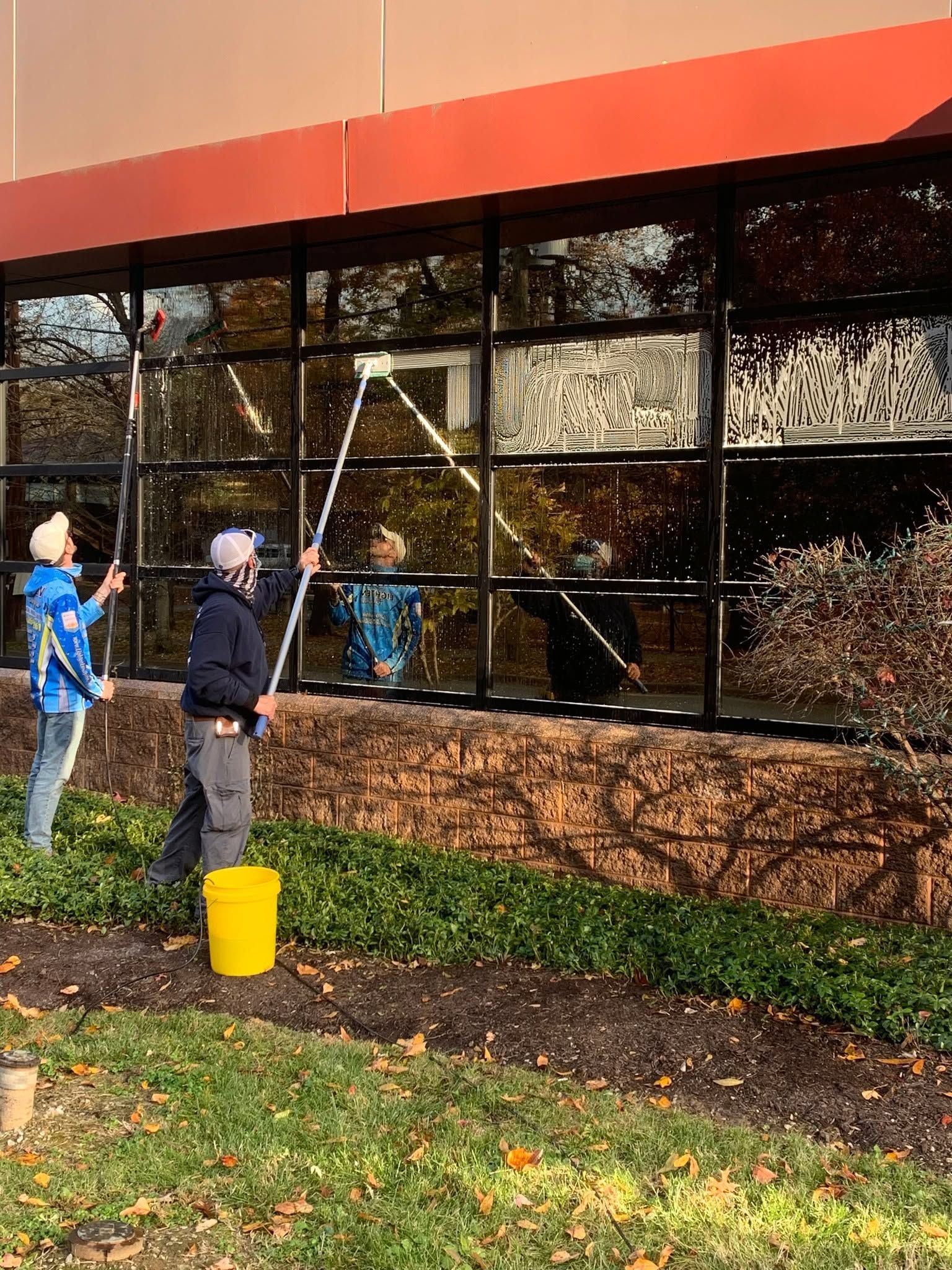 Two people washing large building windows with a long squeegee and a yellow bucket nearby.