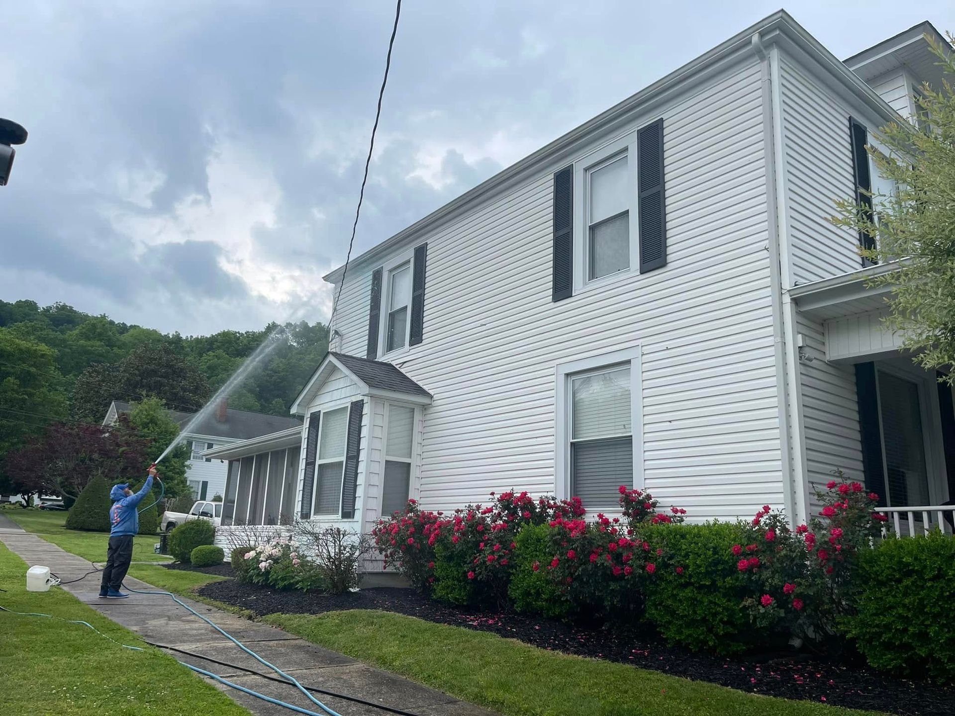 White two-story house with black shutters, red flowers, and a person standing on the sidewalk