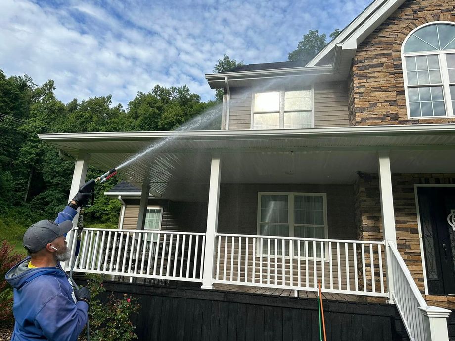Person spraying water onto a two-story house with a pressure washer under a blue sky