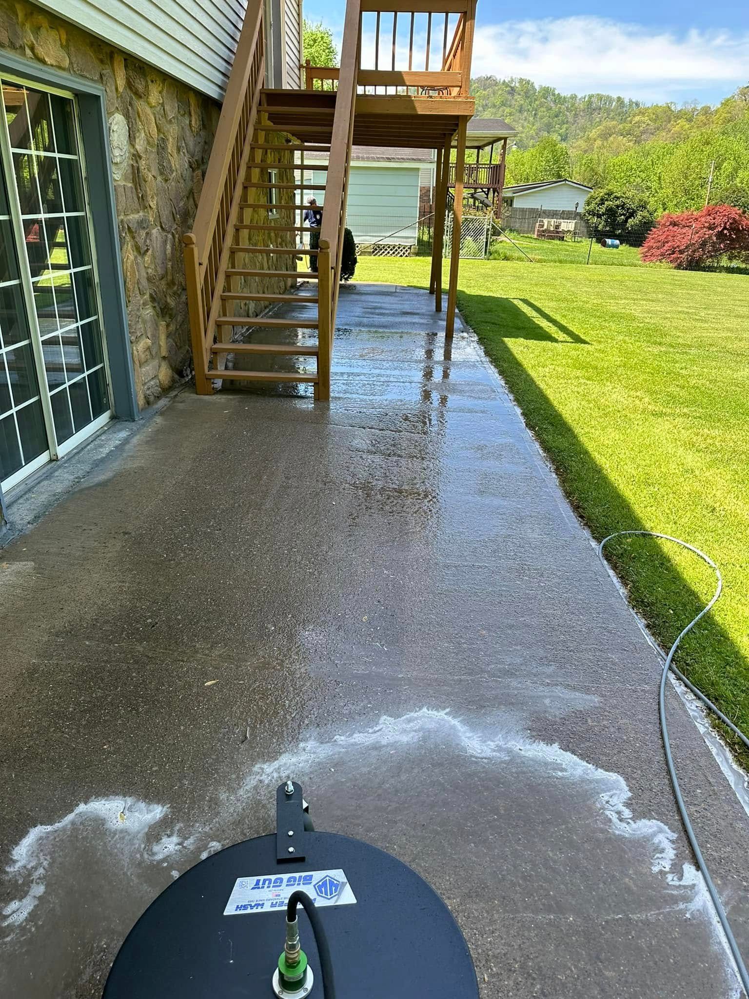 Wet concrete patio beside house stairs with a pressure washer spraying water in the foreground