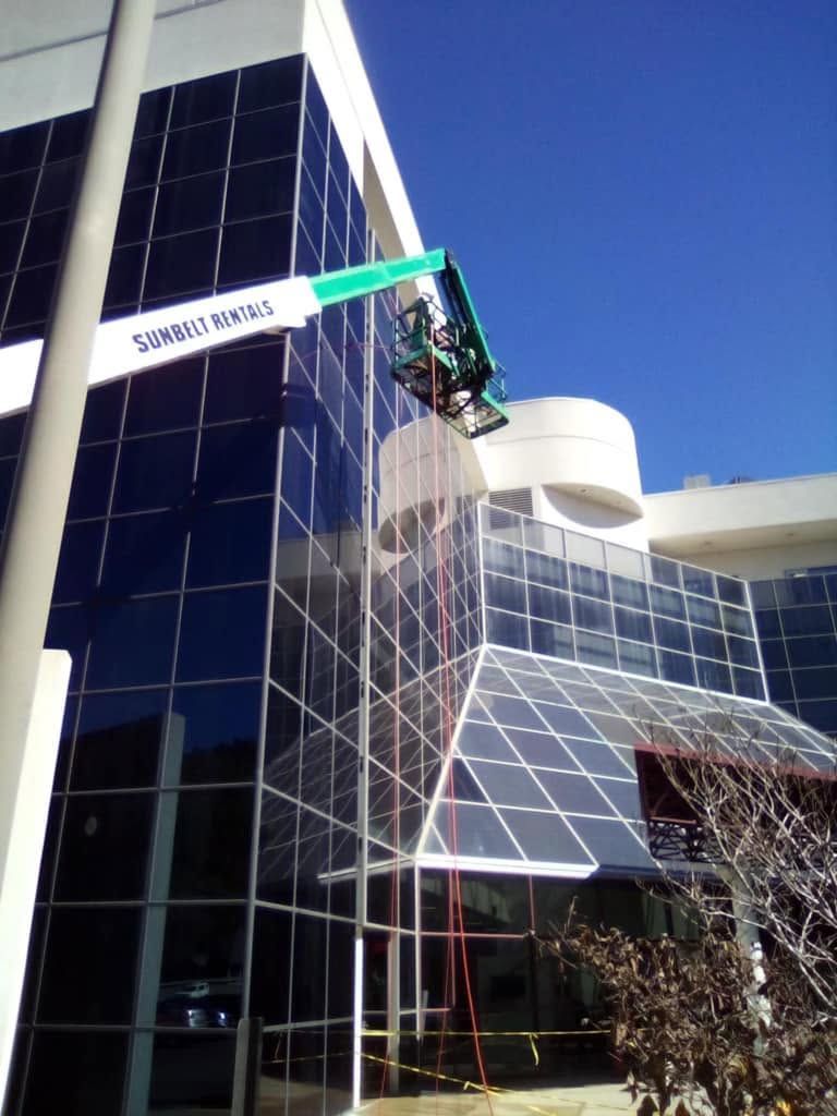 Worker in a green boom lift cleaning windows on a modern glass building under a clear blue sky